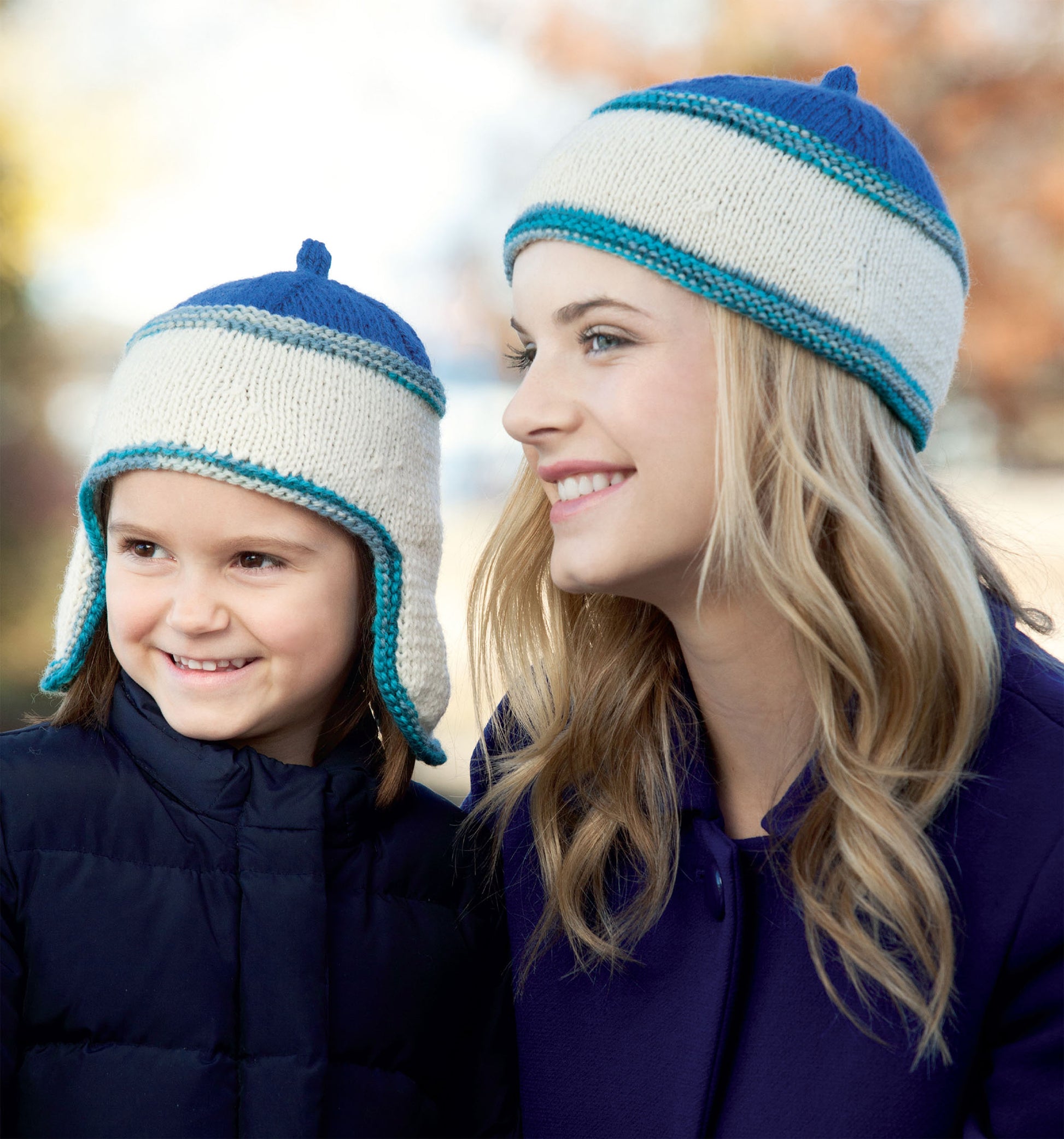 An adult and child wear matching blue and white knit hats with earflaps, smiling outdoors. Create cozy looks like these with the Leisure Arts "Hats and Scarves for Kids" digital download—perfect for beginner knitters. Autumn leaves blur in the background.