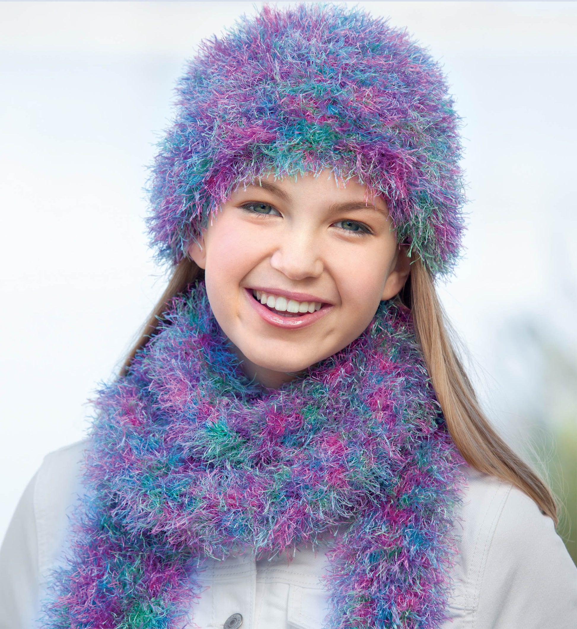 A smiling person in a fluffy, multicolored hat and scarf—ideal for beginner projects—crafted from Leisure Arts' "Hats and Scarves for Kids" digital download, shown in purple, blue, and teal with long hair and a light jacket.