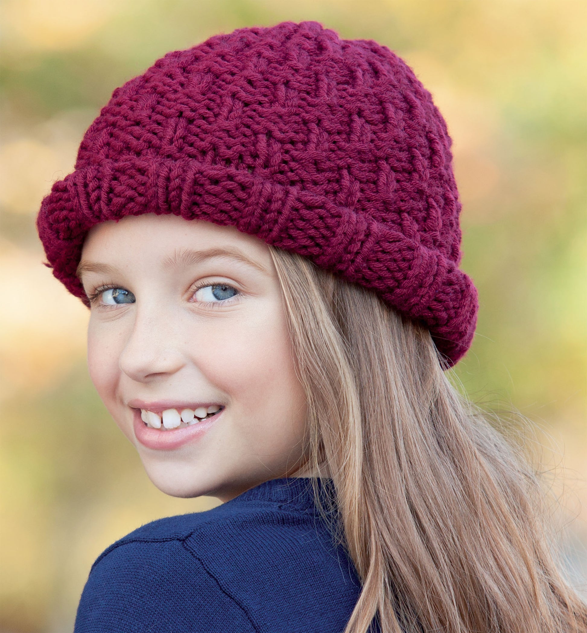 A smiling young girl with long blonde hair wears a deep red knit hat, inspiring beginner projects. Discover similar patterns in the "Hats and Scarves for Kids Digital Download" by Leisure Arts. Warm, inviting outdoor vibes set the scene.
