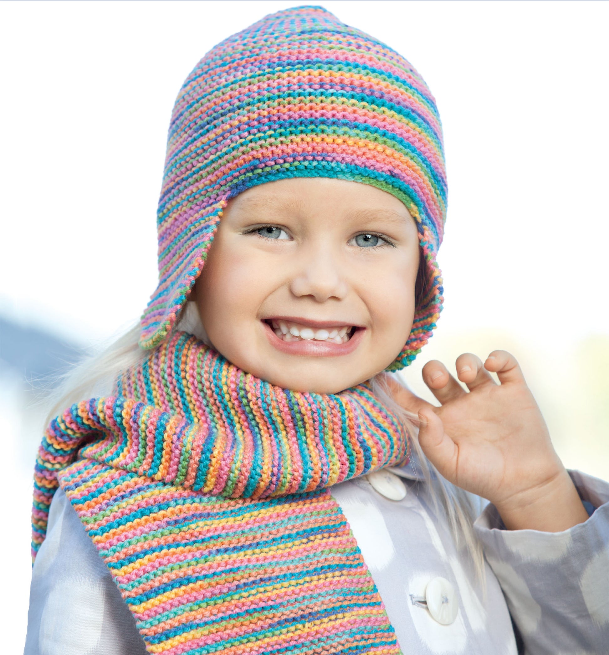 A smiling child in a colorful knit hat and matching striped scarf—made with Leisure Arts’ Hats and Scarves for Kids Digital Download—waves outdoors in a light coat, showcasing the perfect beginner knitting project.