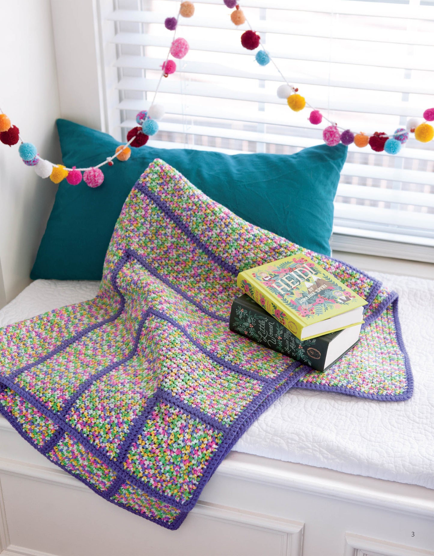 A vibrant crocheted blanket made from the Leisure Arts Easy Afghans Digital Download drapes over a white bench beneath a window with a teal pillow. Three books sit atop the blanket, and a pom-pom garland hangs above.