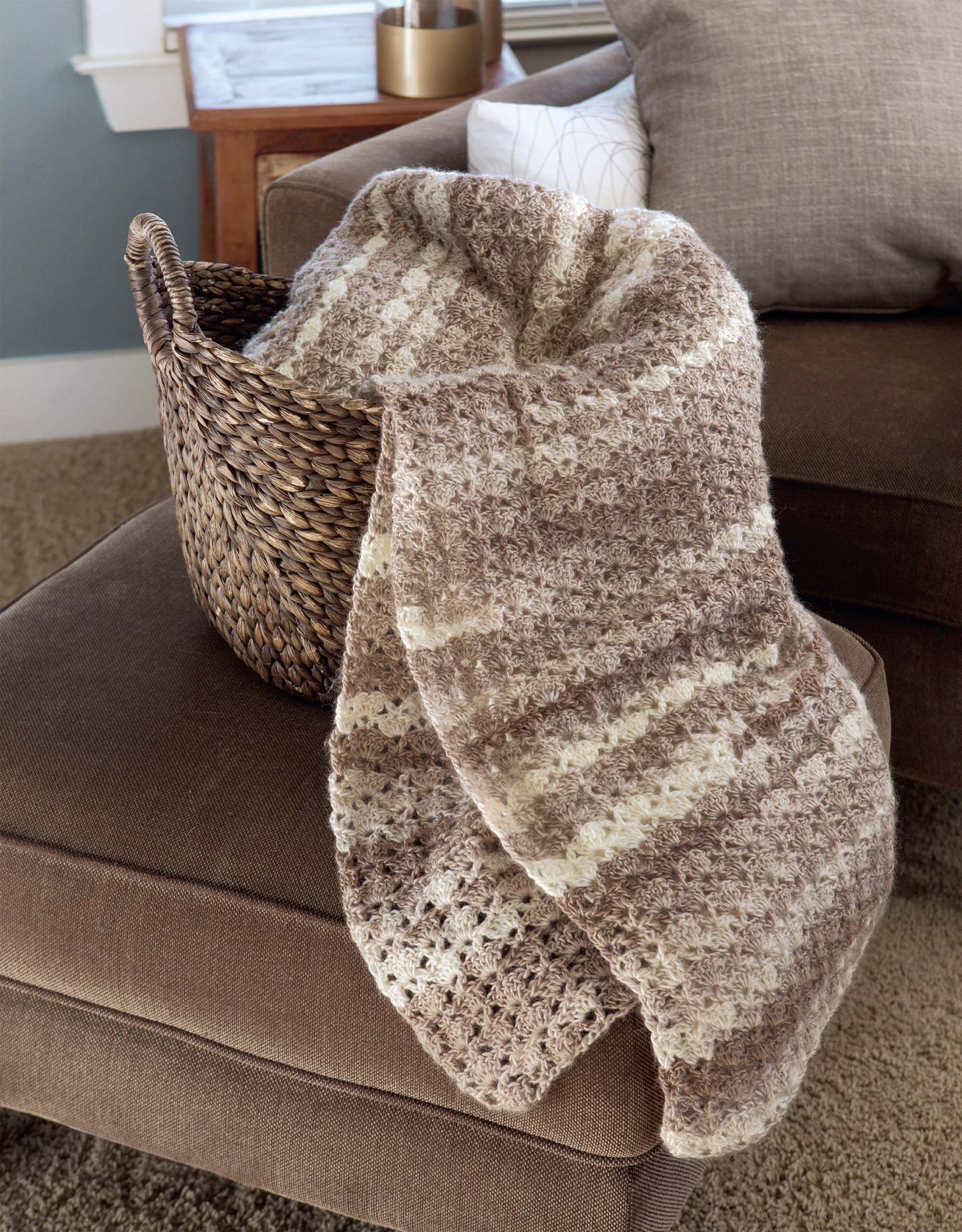 The Easy Afghans Digital Download by Leisure Arts features a cozy, striped crocheted blanket, ideal for beginners. The blanket is shown draped over a basket on a brown ottoman in a stylish living room.