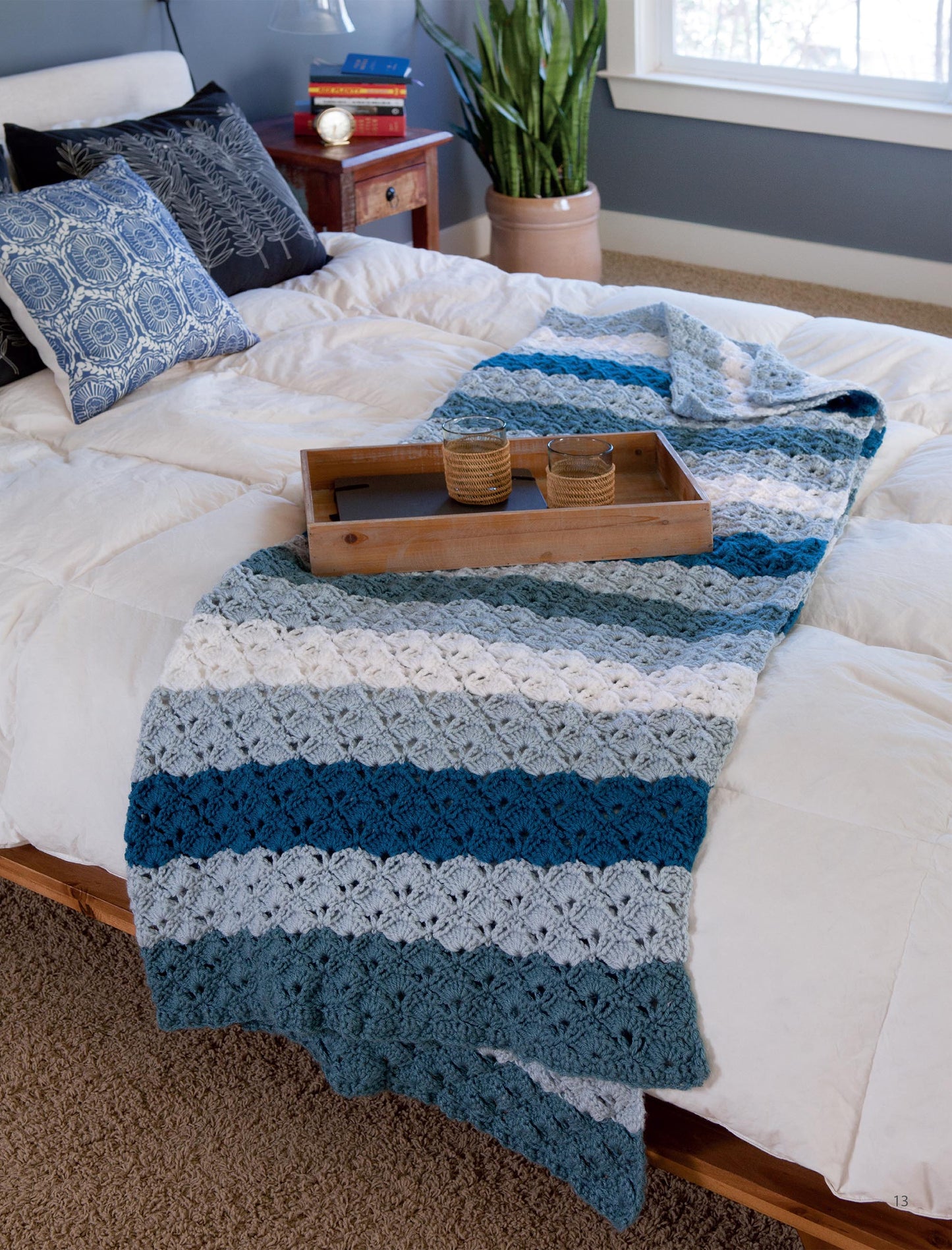 A cozy bedroom features a white bedspread, a blue and white striped blanket made from Leisure Arts’ Easy Afghans Digital Download, a wooden tray with two cups, a potted plant by the window, and decorative pillows on the bed.