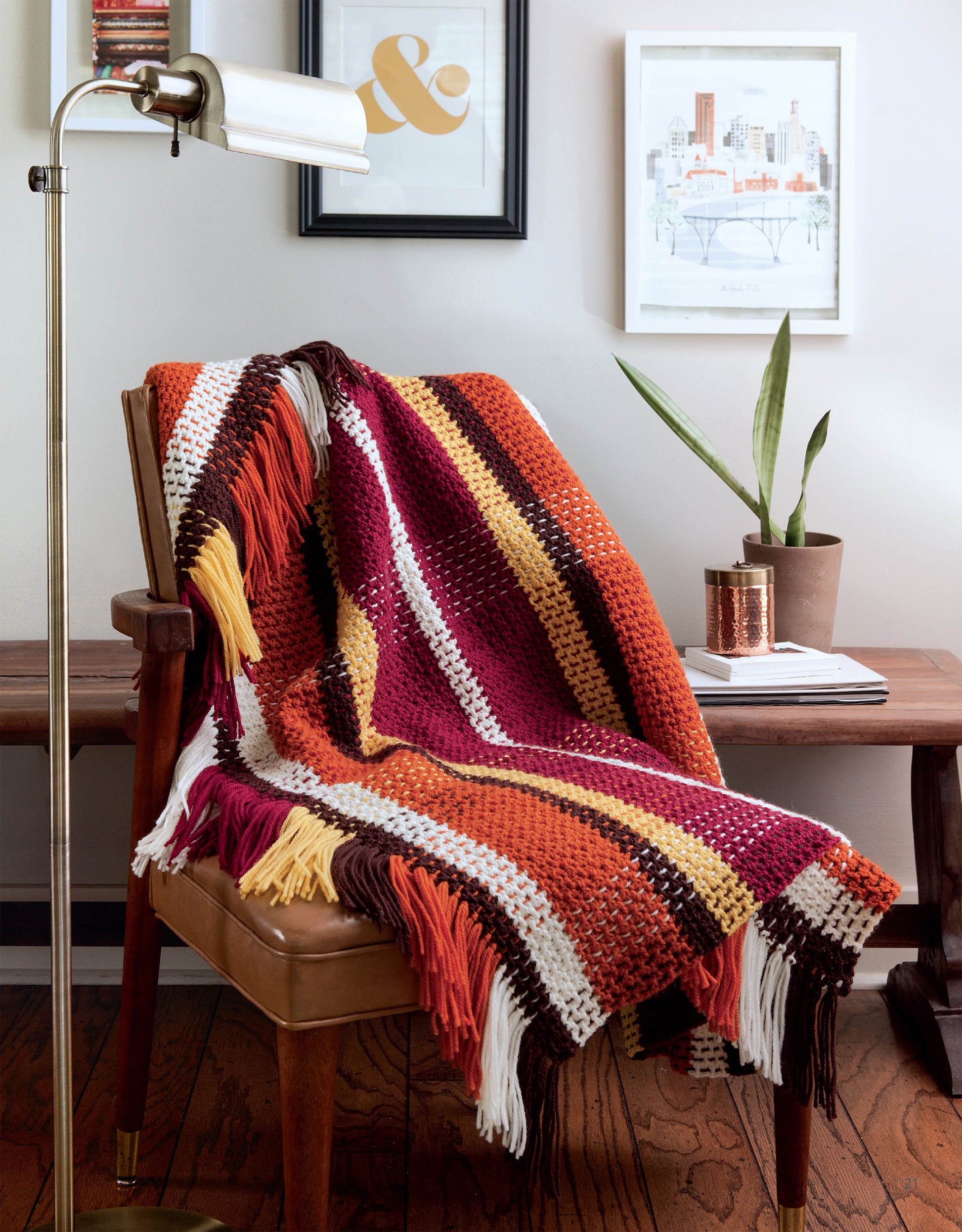 The Easy Afghans Digital Download by Leisure Arts features a multicolored crochet blanket with red, orange, yellow, brown, and white stripes, shown draped over a tan chair beside decor on a wooden floor—great for beginners or easy crochet blankets.