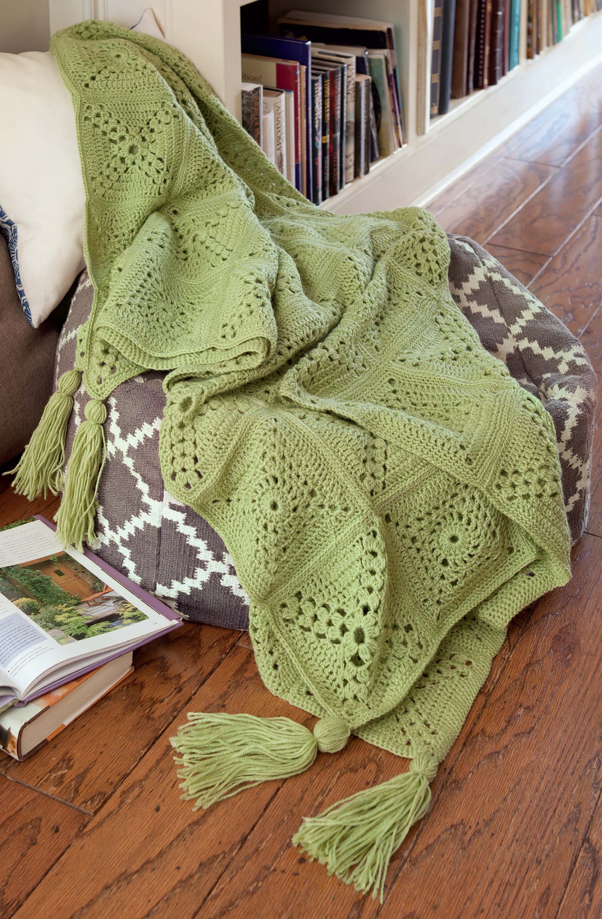 The Easy Afghans Digital Download by Leisure Arts features a green crocheted blanket with square designs and large tassels, draped over a gray-and-white pouf. Books and magazines sit nearby, creating a cozy reading nook.
