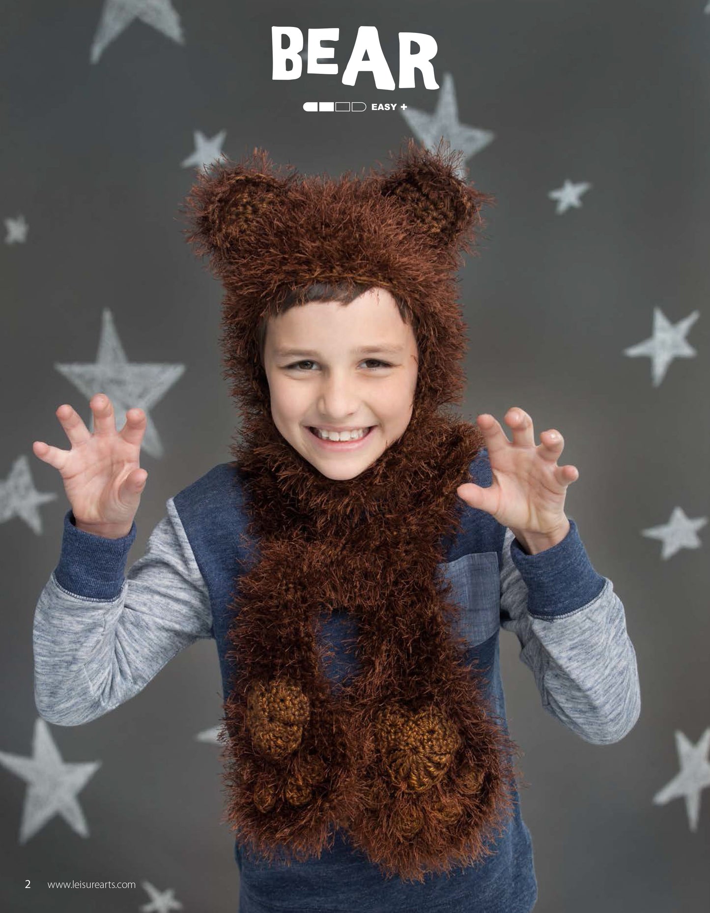 A smiling child wears a fuzzy brown bear hood and scarf made with easy patterns from Animal Hats & Scarves by Leisure Arts, posing with hands raised like bear claws in front of a gray background with white stars and the word BEAR above them.