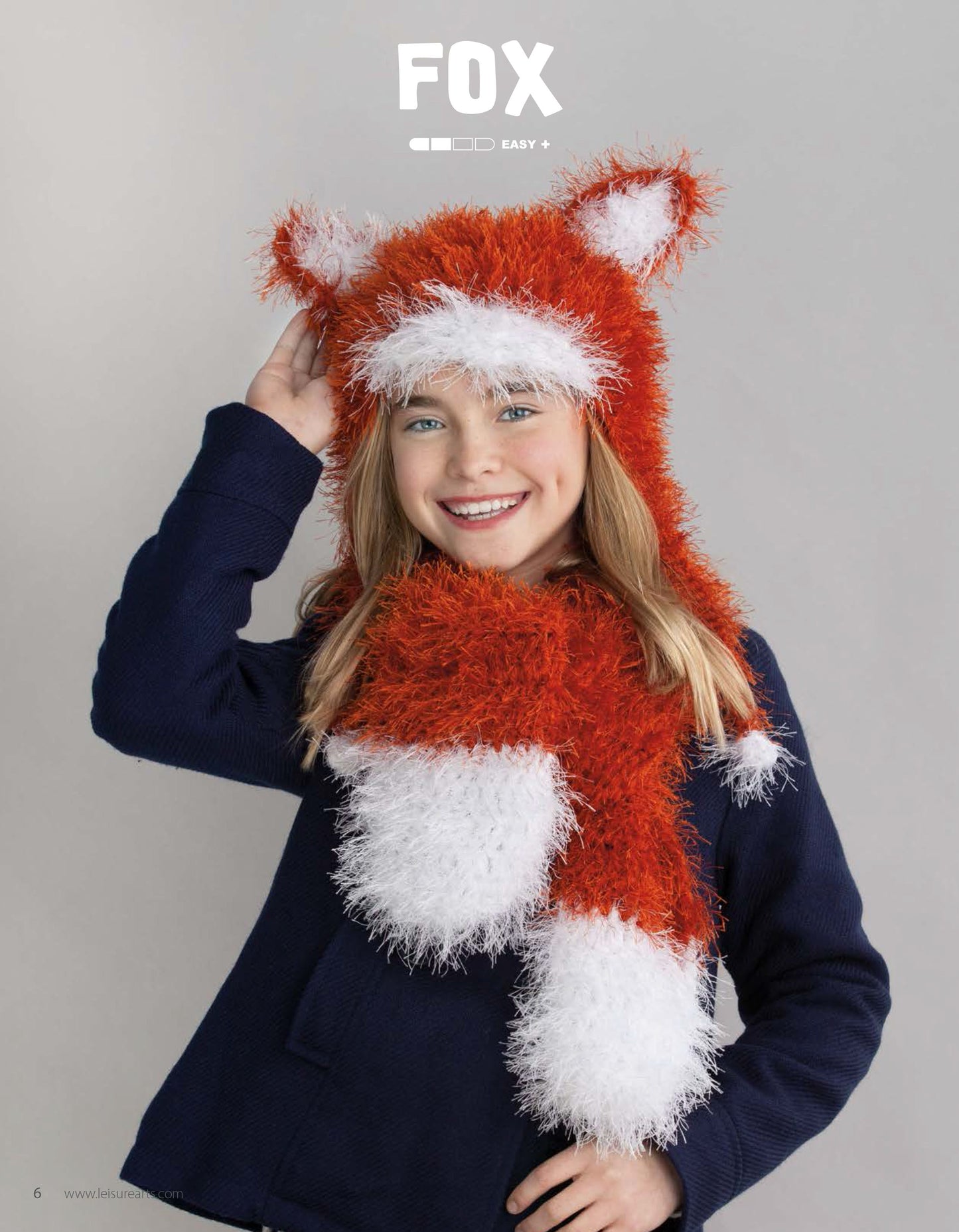 A happy girl wears the Leisure Arts Animal Hats & Scarves set, featuring a cute orange and white fox design with ears and matching scarf, against a plain gray background.
