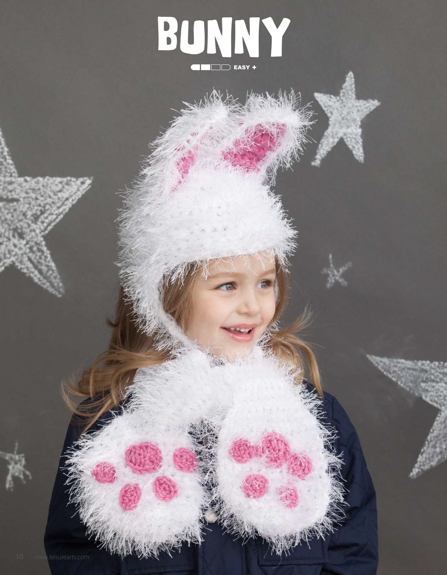 A young child wears Leisure Arts' Animal Hats & Scarves—a fluffy white hat with pink bunny ears, matching mittens, and a cozy scarf—smiling in front of a grey background with white stars.