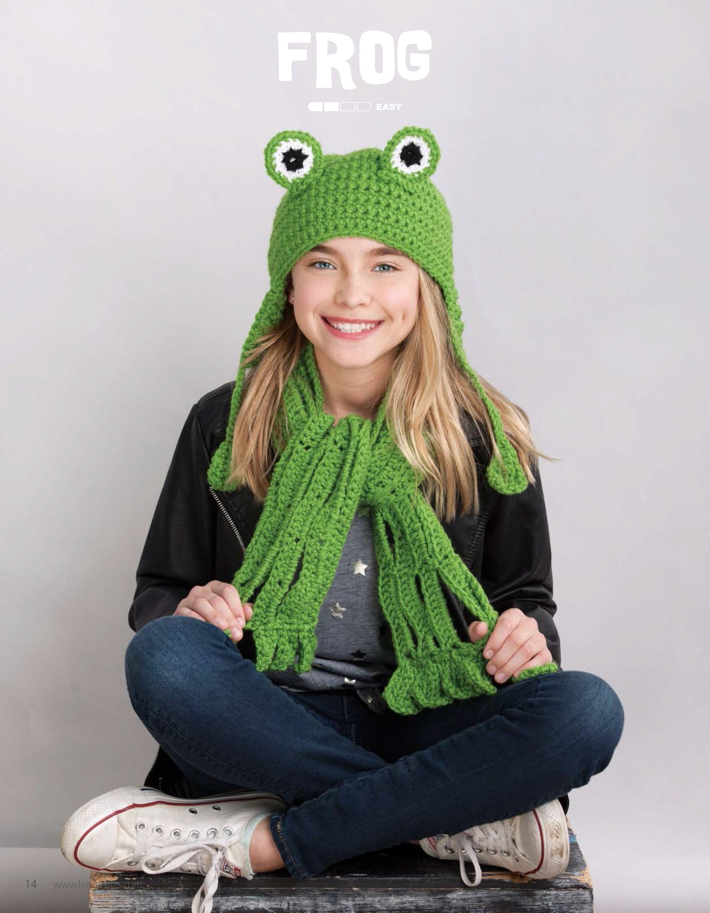 A smiling girl with blonde hair models a green crocheted frog hat and matching scarf from the Leisure Arts Animal Hats & Scarves collection. She sits cross-legged in jeans, a black jacket, and white sneakers against a light gray background.