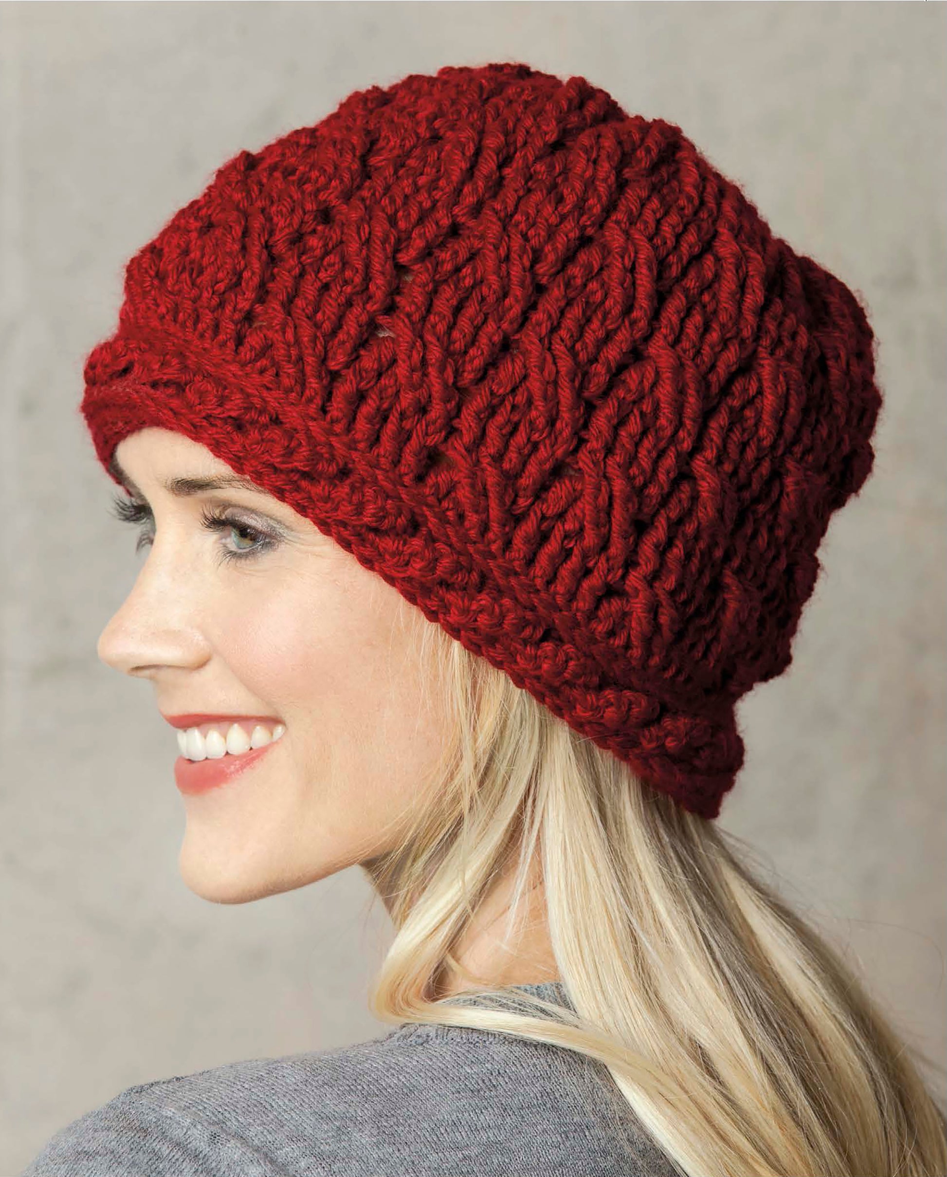 A woman with long blonde hair smiles in profile against a neutral backdrop, wearing a chunky red crochet hat from Leisure Arts' Crochet Caps, Cowls & Mitts collection and a grey top.