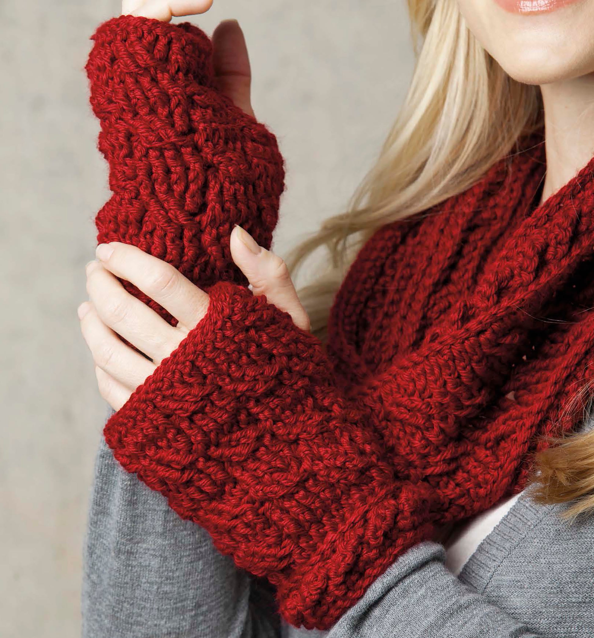 A woman wearing a matching red cabled set of crochet fingerless gloves and an infinity scarf from Crochet Caps, Cowls & Mitts by Leisure Arts, paired with a grey sweater.