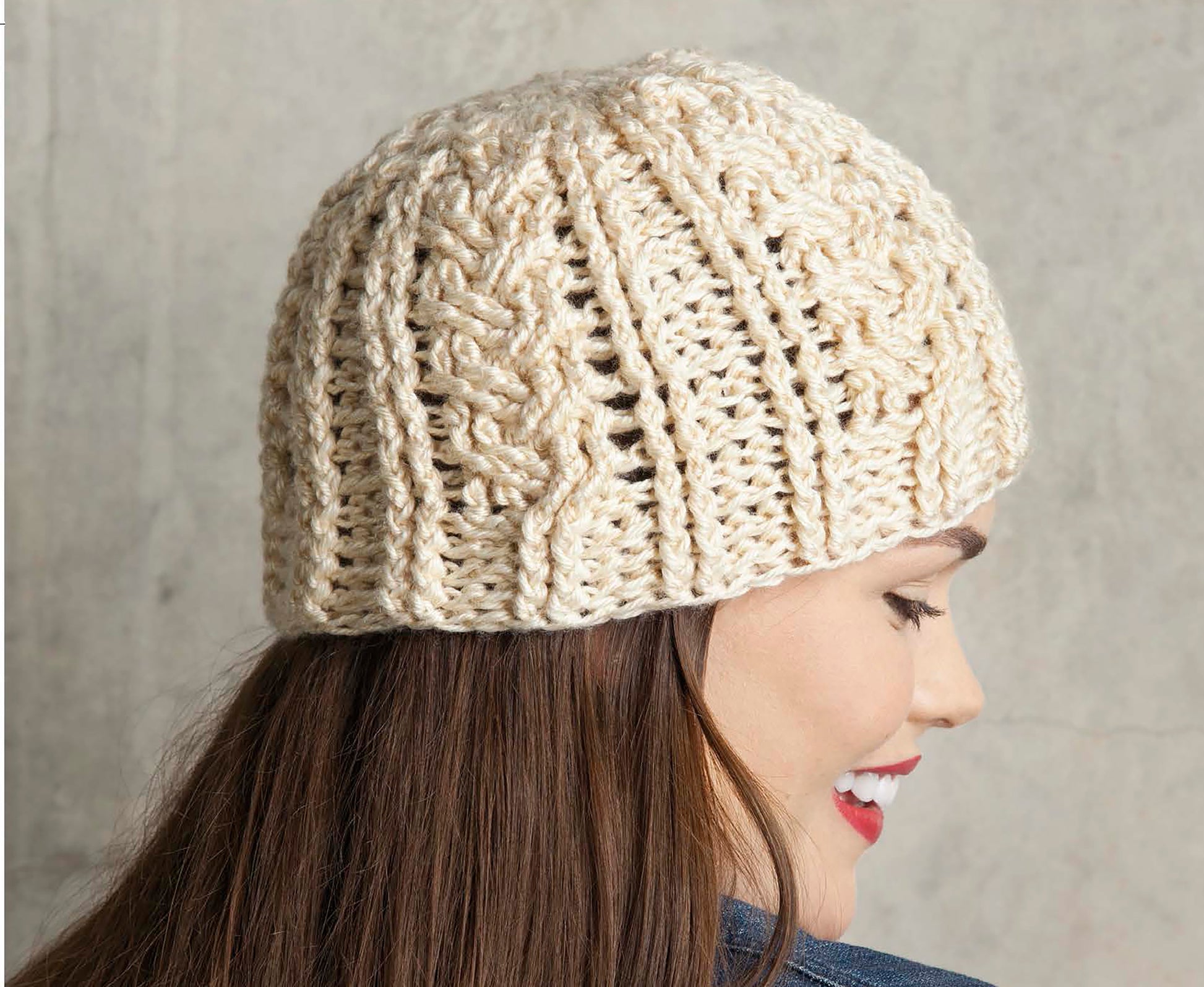 A woman with long brown hair and red lipstick smiles in profile, wearing a chunky cream cable-knit beanie from Leisure Arts' Crochet Caps, Cowls & Mitts, against a plain light background.