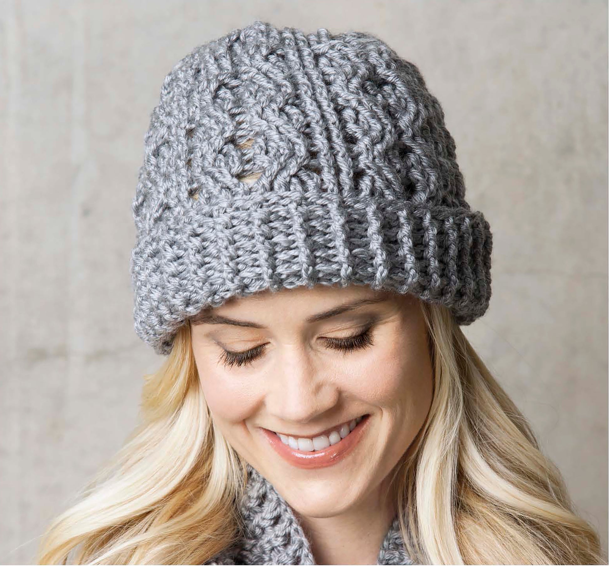 A woman with long blonde hair smiles and looks down while wearing a chunky light gray beanie from the Crochet Caps, Cowls & Mitts collection by Leisure Arts against a neutral background.
