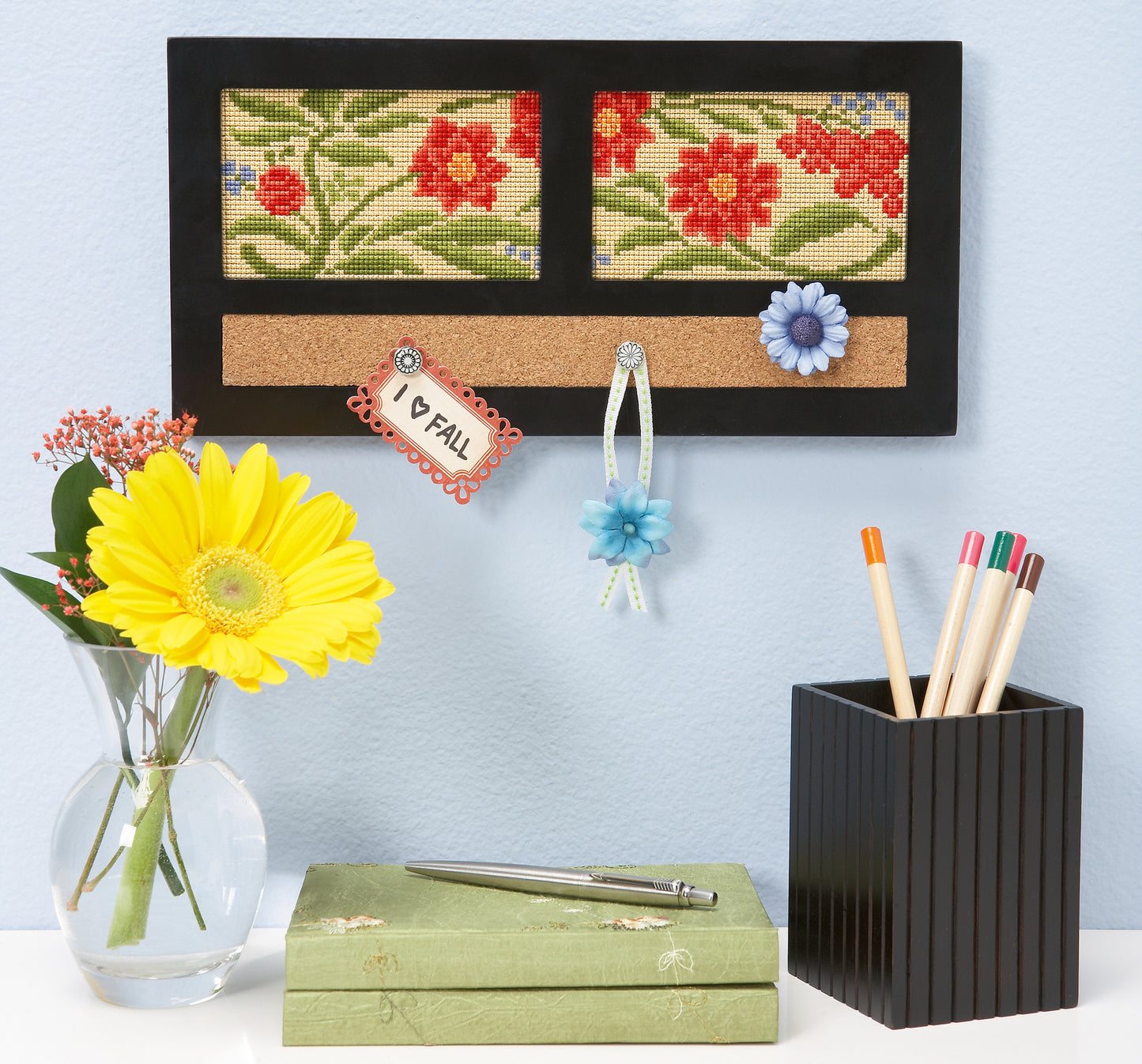 A desk with a vase of yellow gerbera, a green pen box, a black pen holder of colored pens, and a wall organizer decorated with bees and blossoms inspired by the Leisure Arts Fabulous Florals Cross Stitch Digital Download.