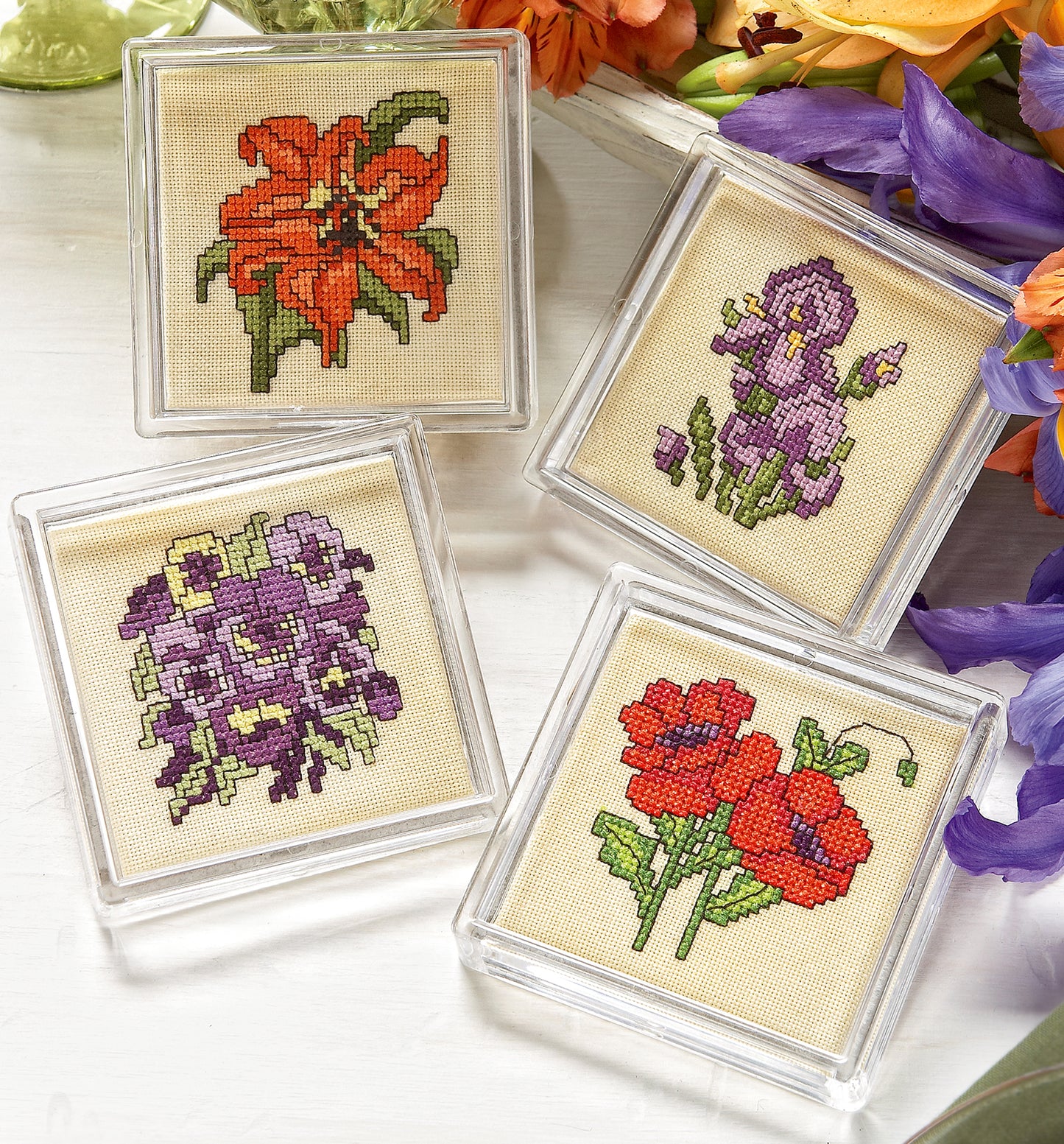 The Fabulous Florals Cross Stitch Digital Download by Leisure Arts features four square coaster designs with a clear frame, each showcasing a bright flower—orange lily, purple iris, violet pansies, red poppies—and hints of bees for extra charm.