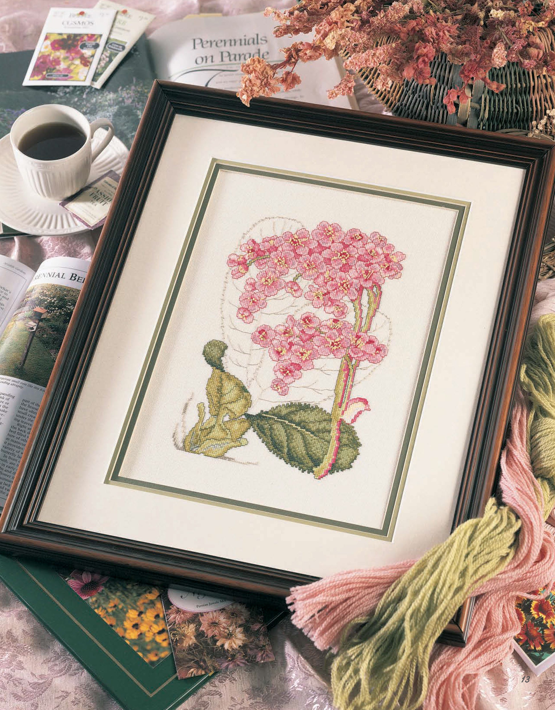 The Fabulous Florals Cross Stitch Digital Download by Leisure Arts is displayed framed with gardening books, embroidery floss, coffee, and a pink bouquet arranged on the table.