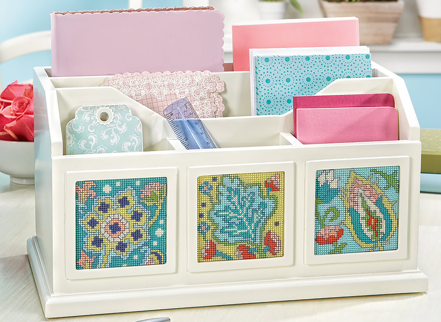 The Fabulous Florals Cross Stitch Digital Download by Leisure Arts is displayed on a light surface, accompanied by pink envelopes, lace, a ruler, patterned tags, and craft papers. The design features blue florals inspired by bees and blossoms.