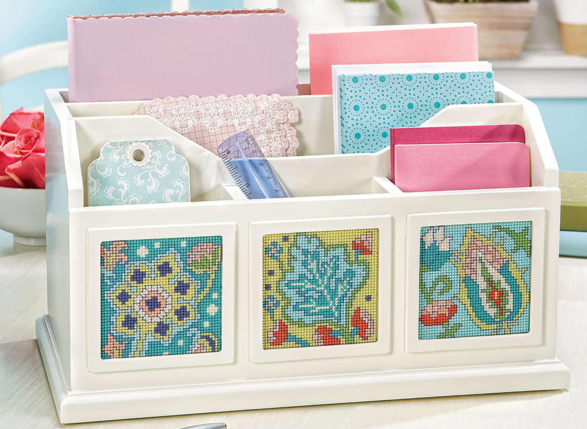 The Fabulous Florals Cross Stitch Digital Download by Leisure Arts is displayed on a light surface, accompanied by pink envelopes, lace, a ruler, patterned tags, and craft papers. The design features blue florals inspired by bees and blossoms.