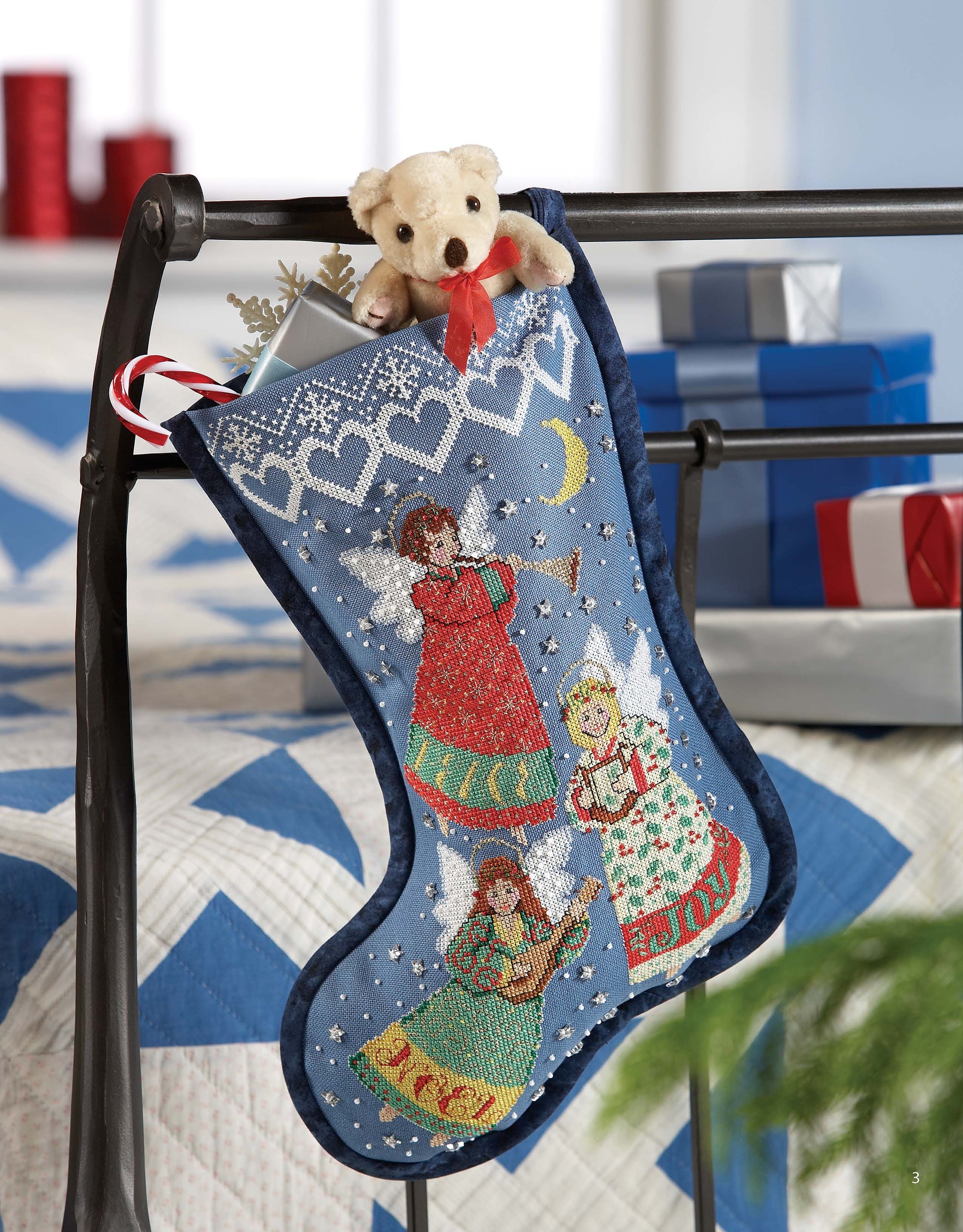 A Festive Christmas Stocking by Leisure Arts, featuring three embroidered angels in blue, hangs on a metal bed frame. The stocking holds a teddy bear, candy cane, and snowflake ornament; wrapped gifts and a quilted bed appear in the background.