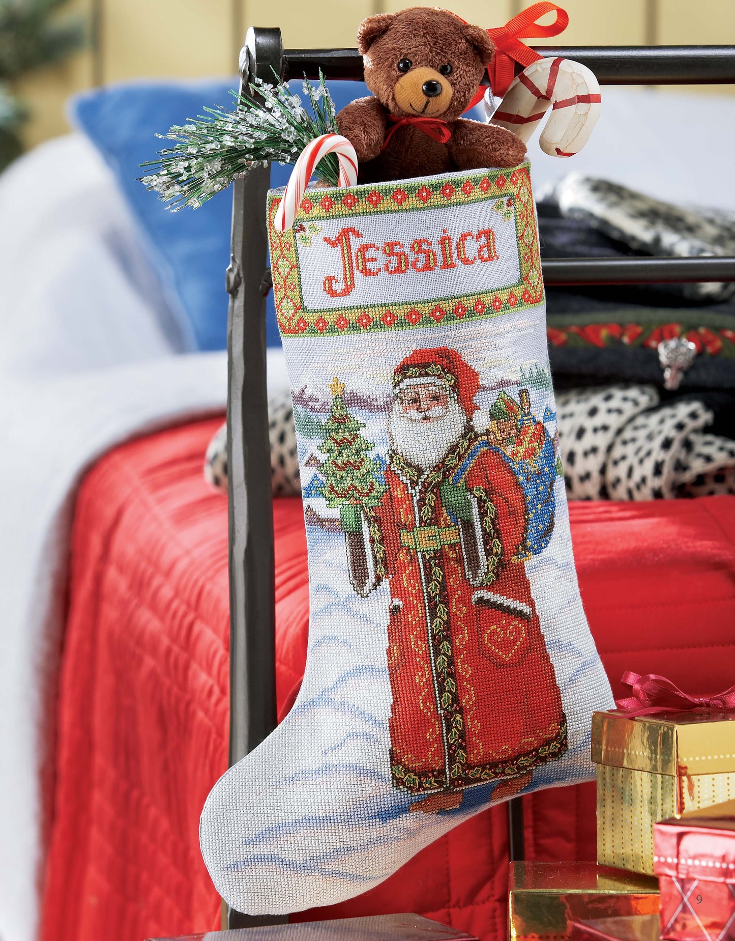 A Festive Christmas Stocking by Leisure Arts hangs on a bed, displaying "Jessica," Santa, and classic holiday designs. Filled with a teddy bear, candy canes, greenery, and gifts, this stocking is the perfect festive touch.