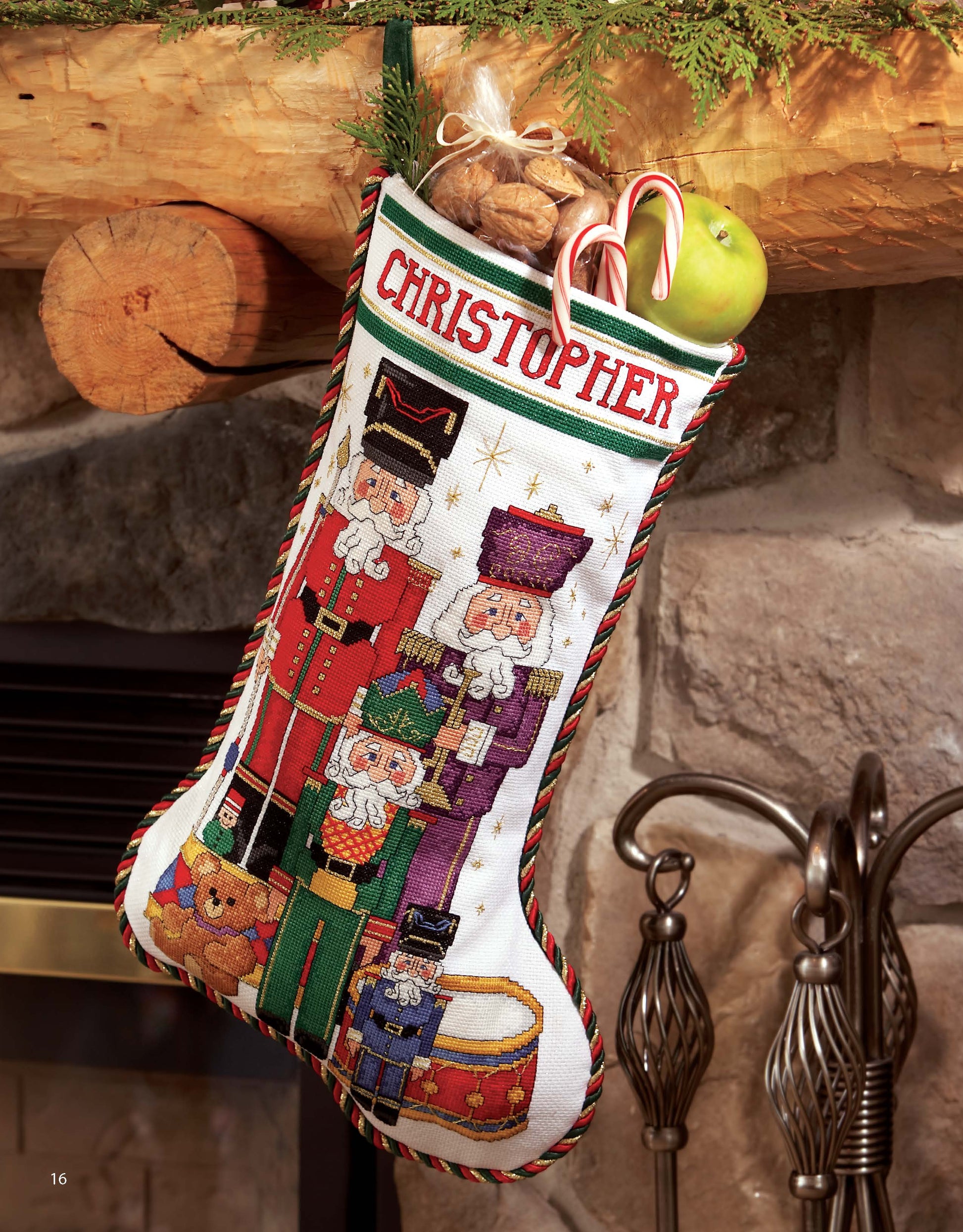 A Leisure Arts Festive Christmas Stocking labeled Christopher hangs on a stone fireplace, embroidered with nutcrackers and holiday motifs, filled with walnuts, candy canes, and a green apple; fir branches trim the mantel above.