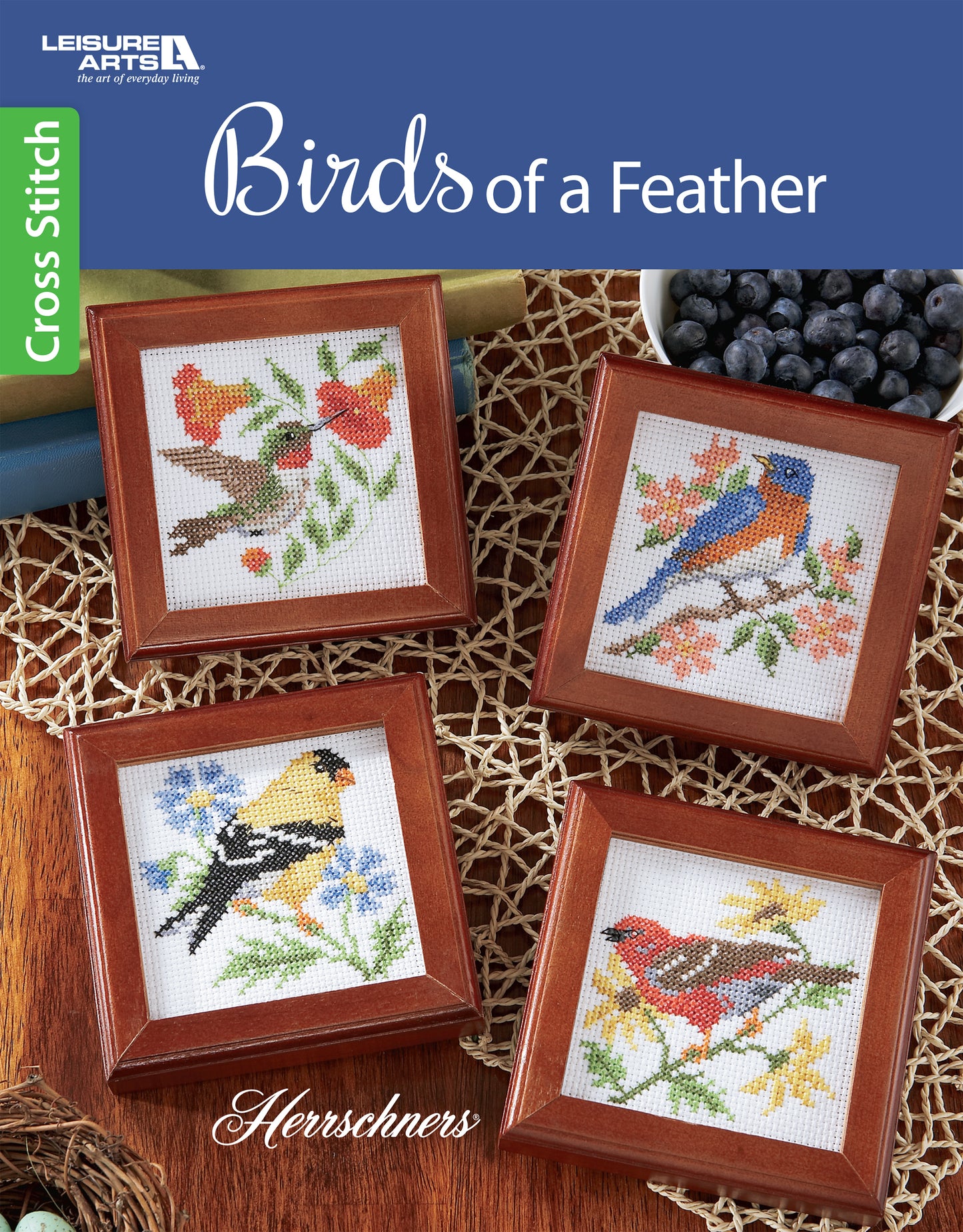 Four framed bird cross stitch patterns from Leisure Arts' Birds of A Feather Cross Stitch feature colorful birds on branches and the title at the top, with nearby blueberries and coasters completing the charming embroidery display.