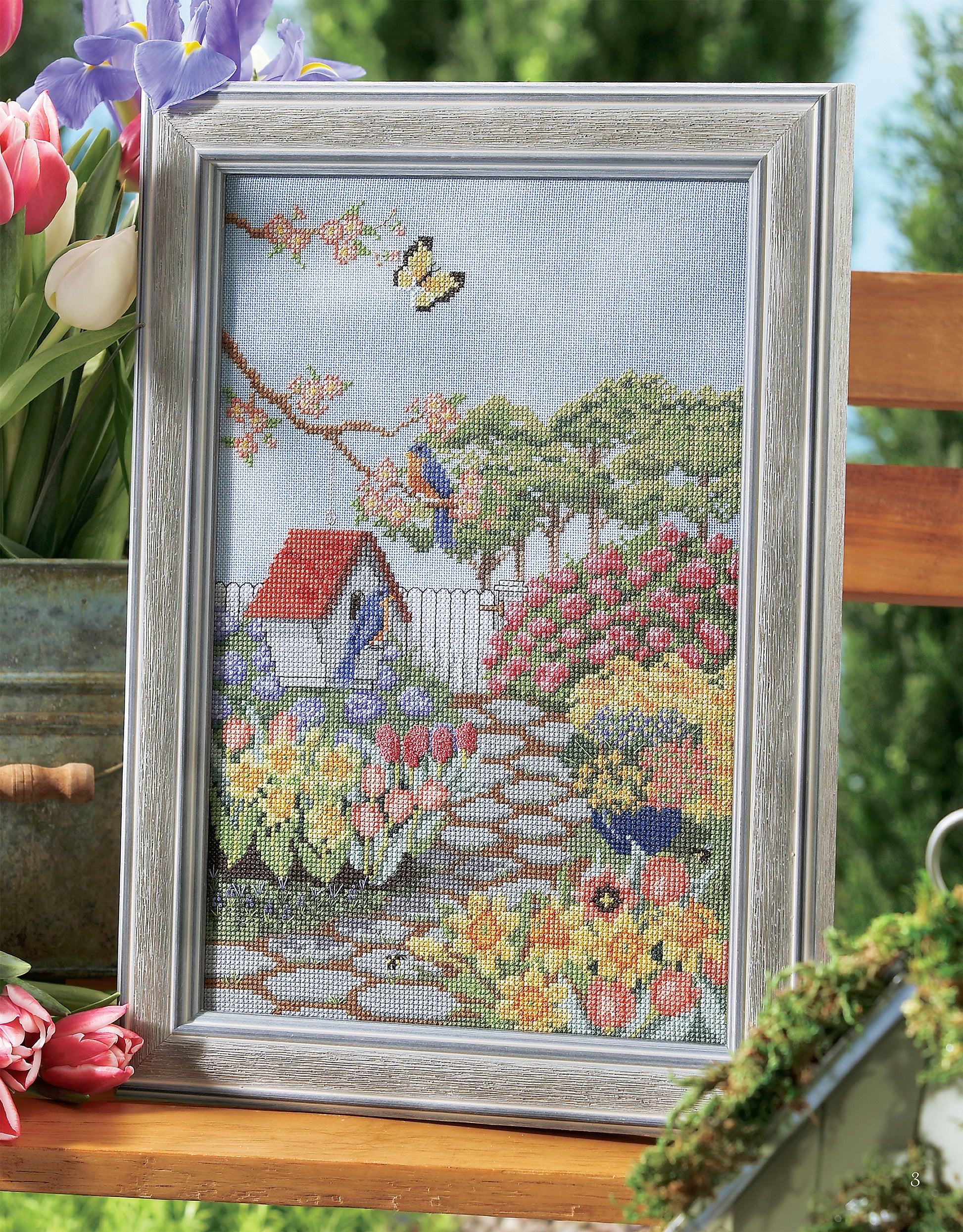 A framed Leisure Arts Birds of A Feather Cross Stitch features a colorful garden scene with flowers, a birdhouse, birds on a branch, and a white picket fence, displayed outdoors on a wooden bench with fresh flowers nearby.