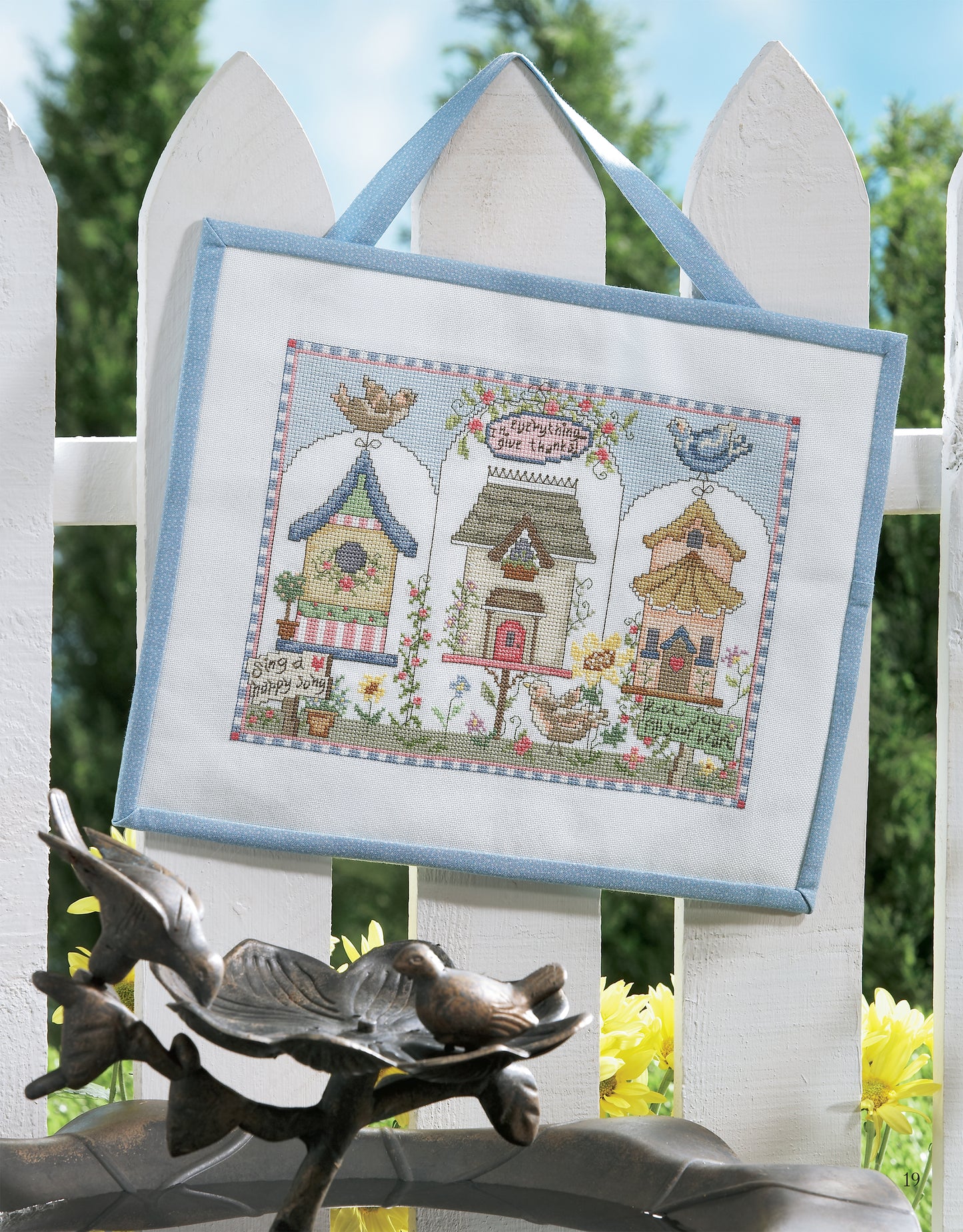 The Birds of A Feather Cross Stitch by Leisure Arts features three birdhouses and birds on a white picket fence, with yellow flowers and green trees against a blue sky—a charming embroidery for nature lovers.