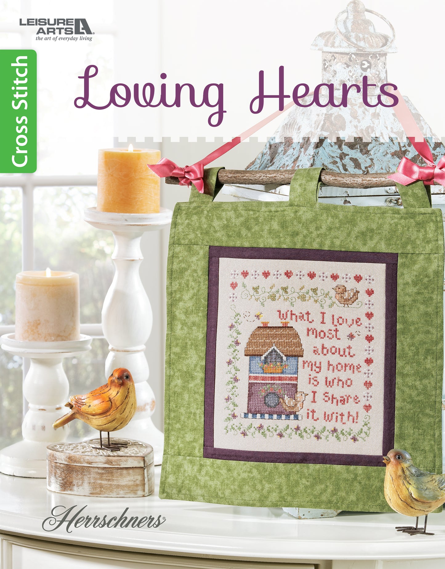 The Leisure Arts Loving Hearts Cross Stitch kit features a decorative design with a house, hearts, and flowers on green fabric, plus the phrase "What I love most about my home is who I share it with.