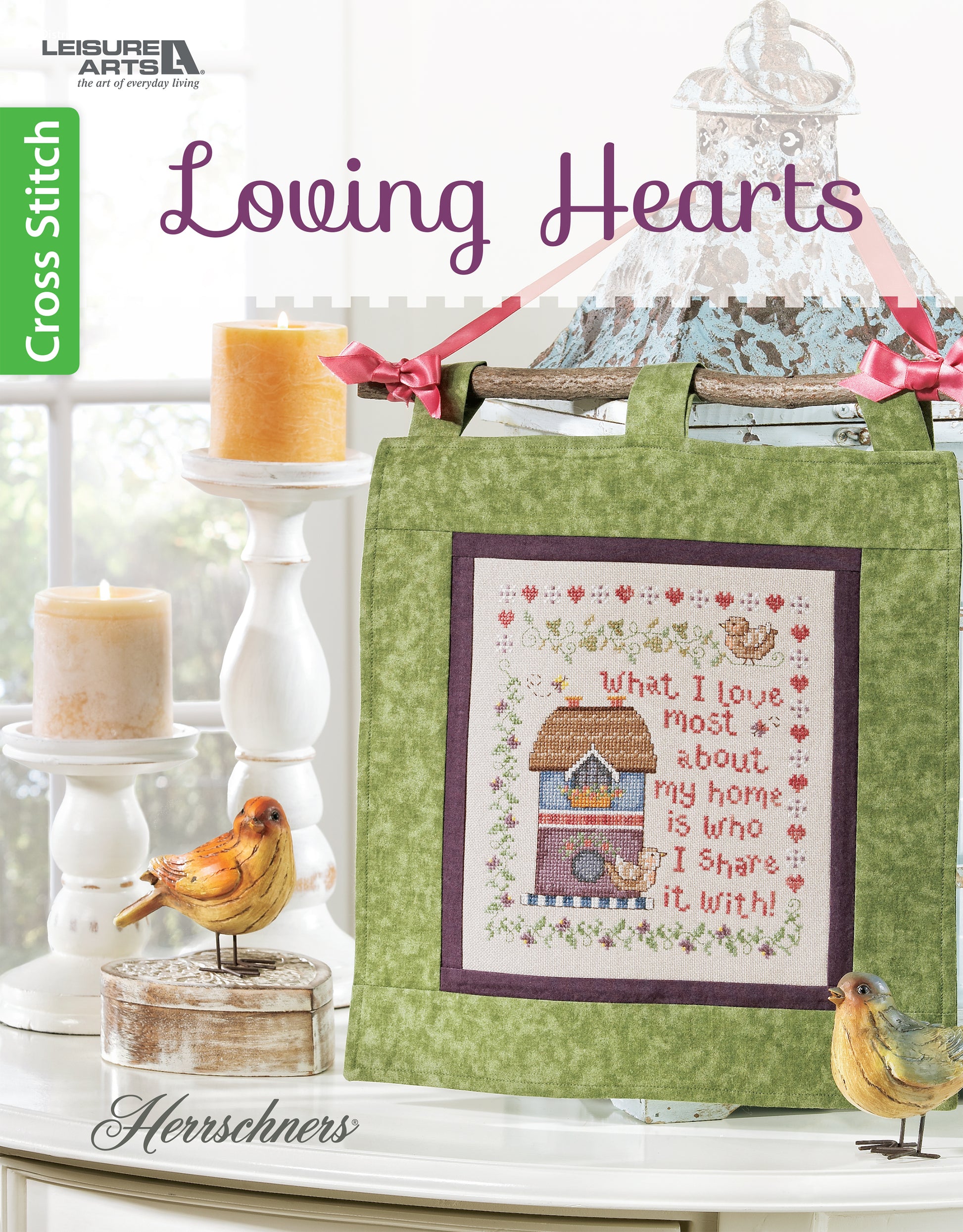 The Leisure Arts Loving Hearts Cross Stitch kit features a decorative design with a house, hearts, and flowers on green fabric, plus the phrase "What I love most about my home is who I share it with.