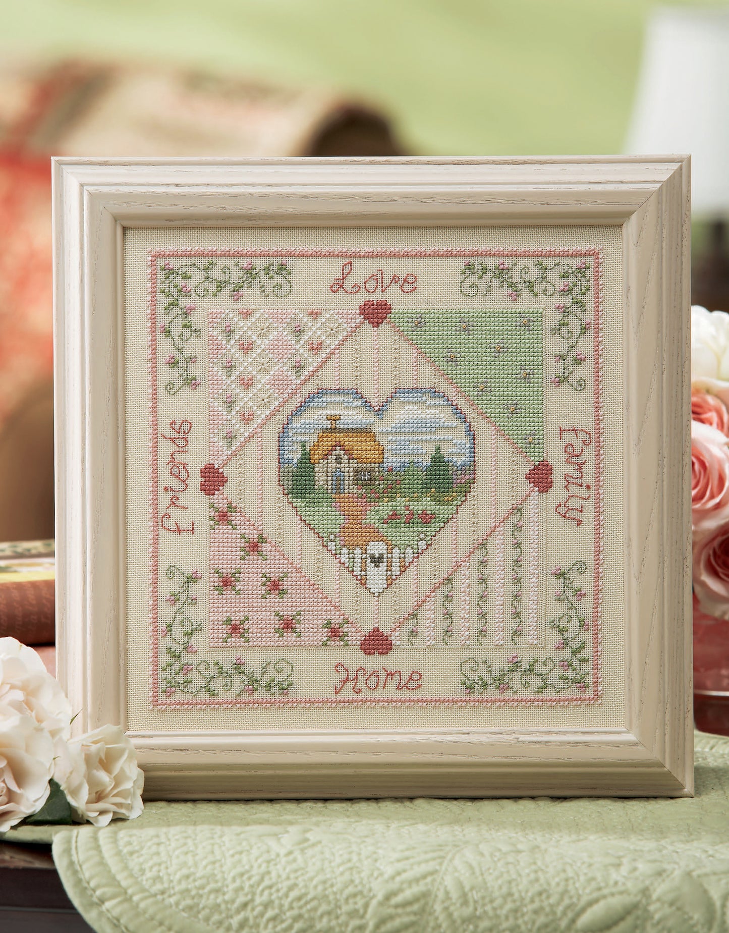 The Leisure Arts Loving Hearts Cross Stitch is a framed artwork with a central cottage, floral accents, and “Love,” “Family,” “Home,” and “Friends” in pink—an ideal, charming decor piece for any home.
