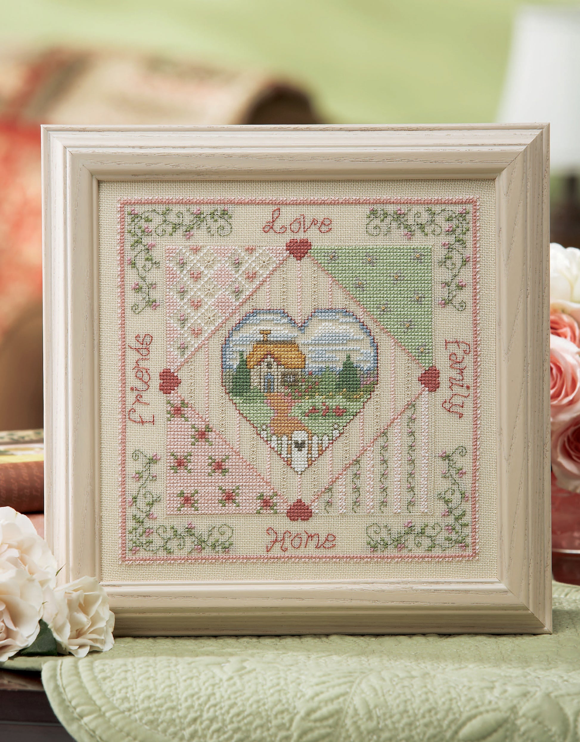 The Leisure Arts Loving Hearts Cross Stitch is a framed artwork with a central cottage, floral accents, and “Love,” “Family,” “Home,” and “Friends” in pink—an ideal, charming decor piece for any home.