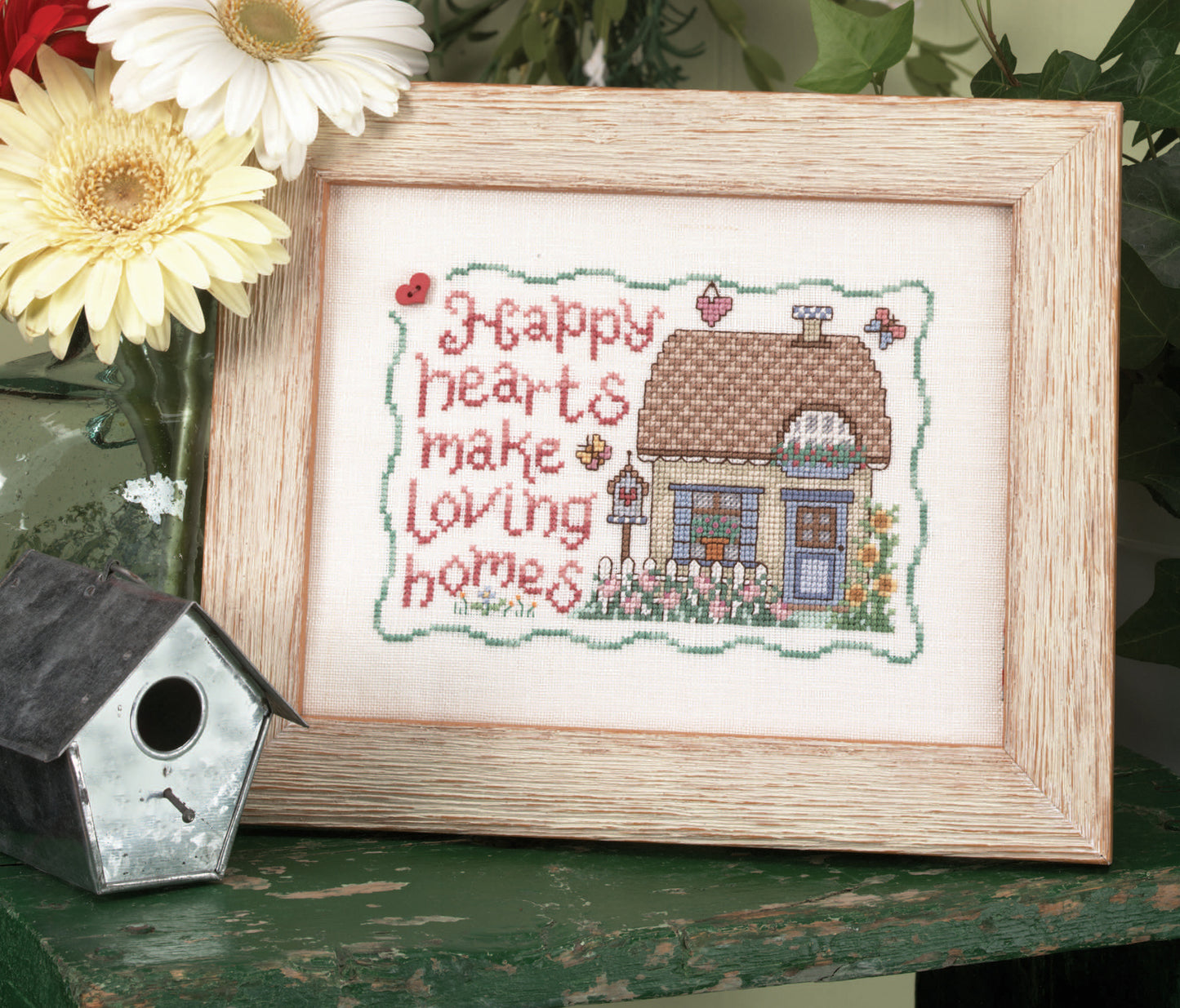 The Loving Hearts Cross Stitch by Leisure Arts features a house, flowers, and “Happy hearts make loving homes,” bordered in green with a birdhouse and daisies—an ideal piece for unique home decor.