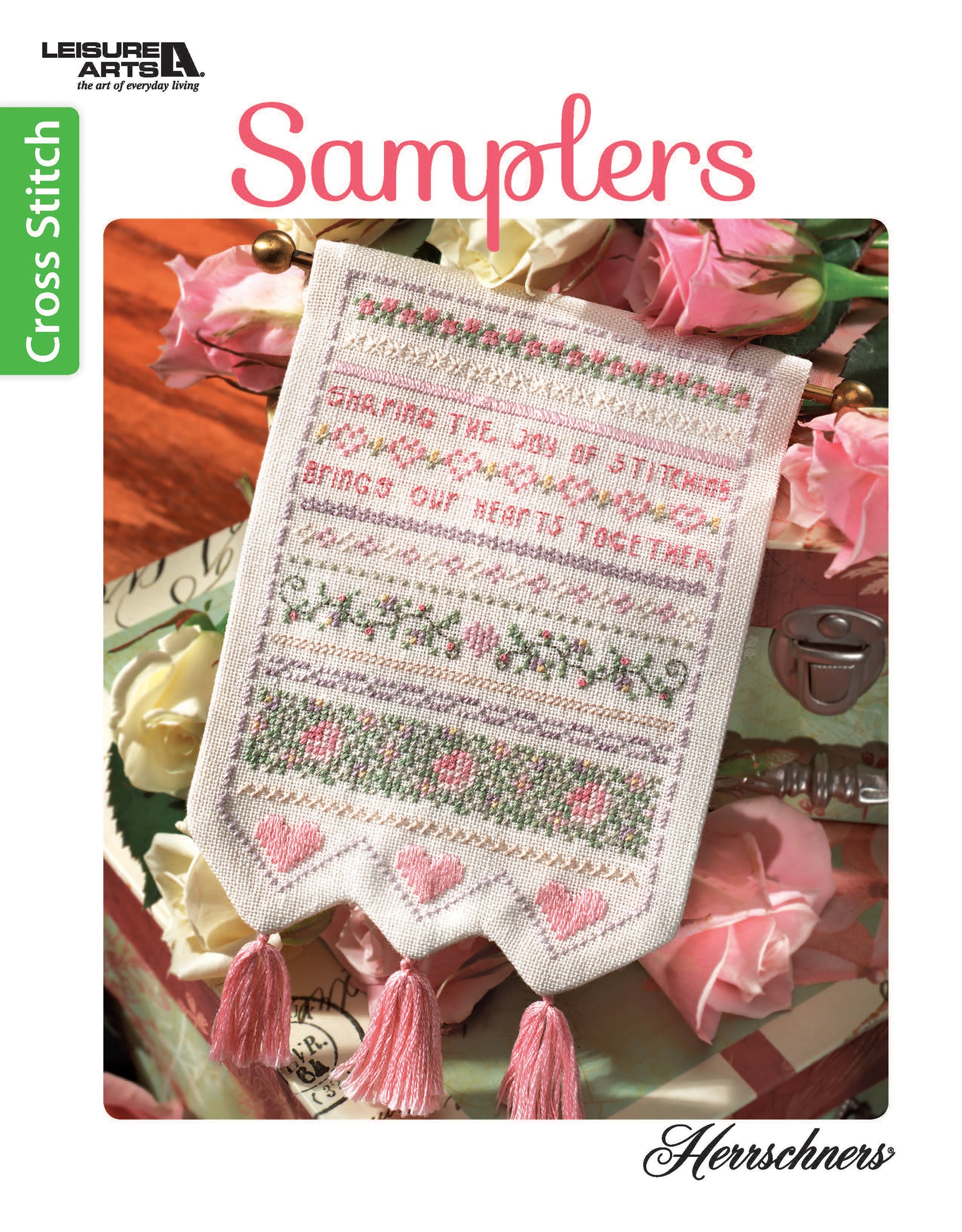 The cover of Leisure Arts' Samplers Cross Stitch booklet features a decorative sampler with pink and green patterns, hearts, and tassels, displayed among pink roses.