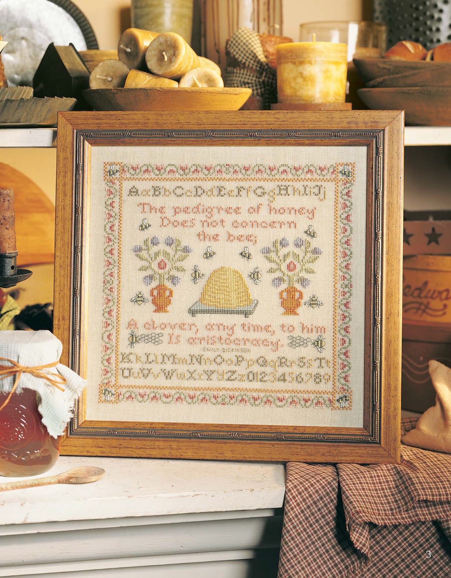 The Samplers Cross Stitch by Leisure Arts, featuring an alphabet, beehive, flowers, and honey-themed text in a rustic frame, adds cozy country charm to shelves with candles and pottery—great inspiration for sampler designs.
