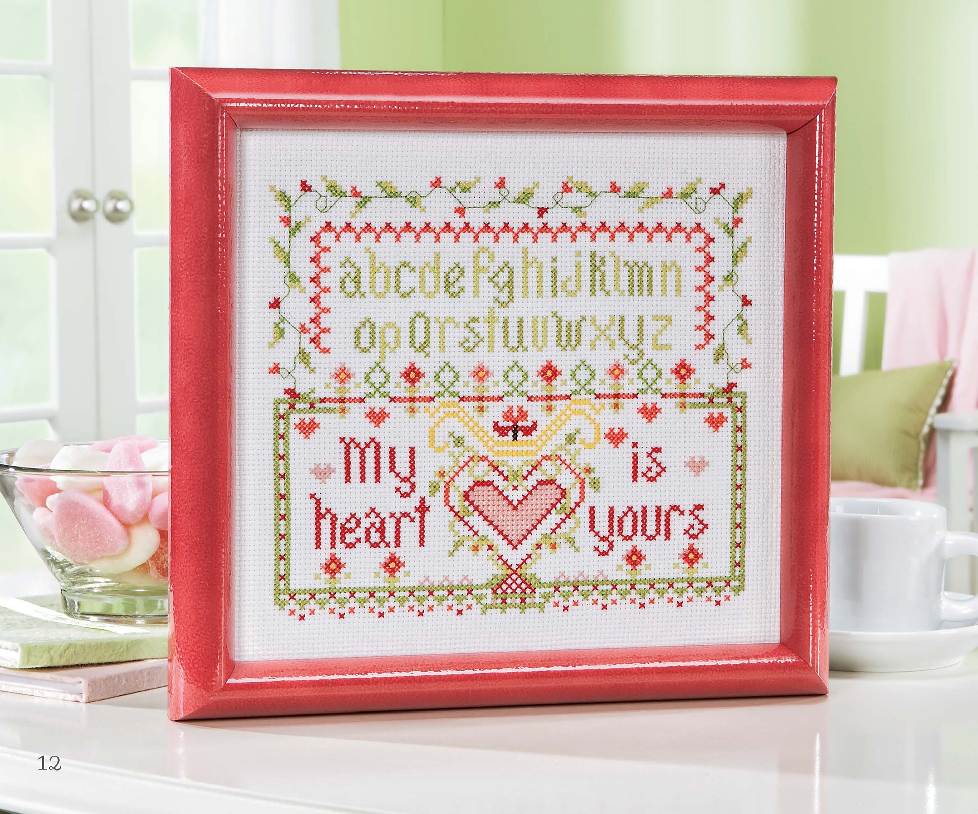 The Leisure Arts Samplers Cross Stitch, featuring a framed green alphabet and "My heart is yours" in red, is displayed on a white table with a cup, saucer, and a bowl of pink and white marshmallows nearby.