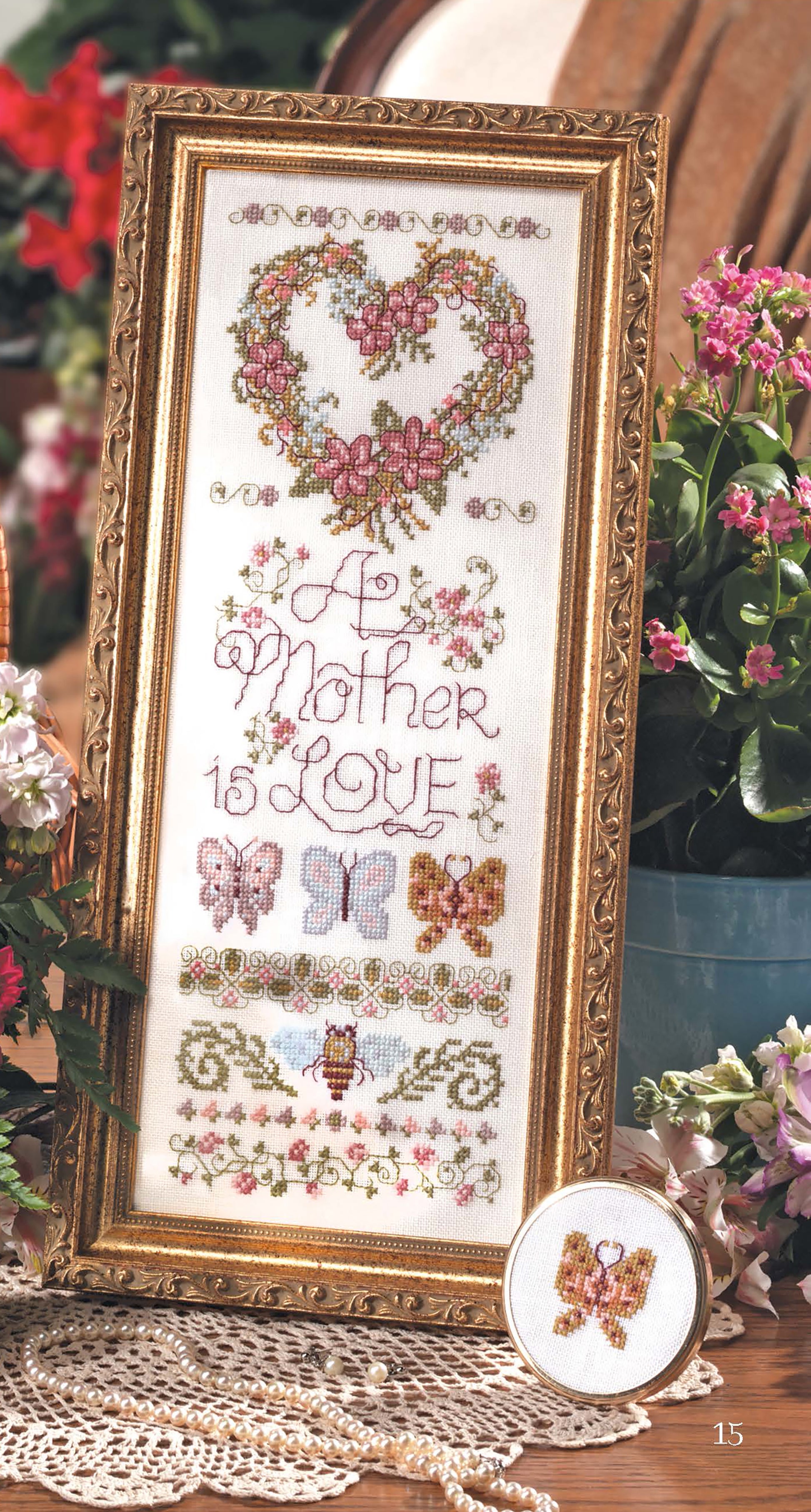 A Leisure Arts Samplers Cross Stitch piece, framed and featuring a heart, flowers, butterflies, and "A Mother is Love," sits on a lace-covered table next to a small round-framed butterfly embroidery.