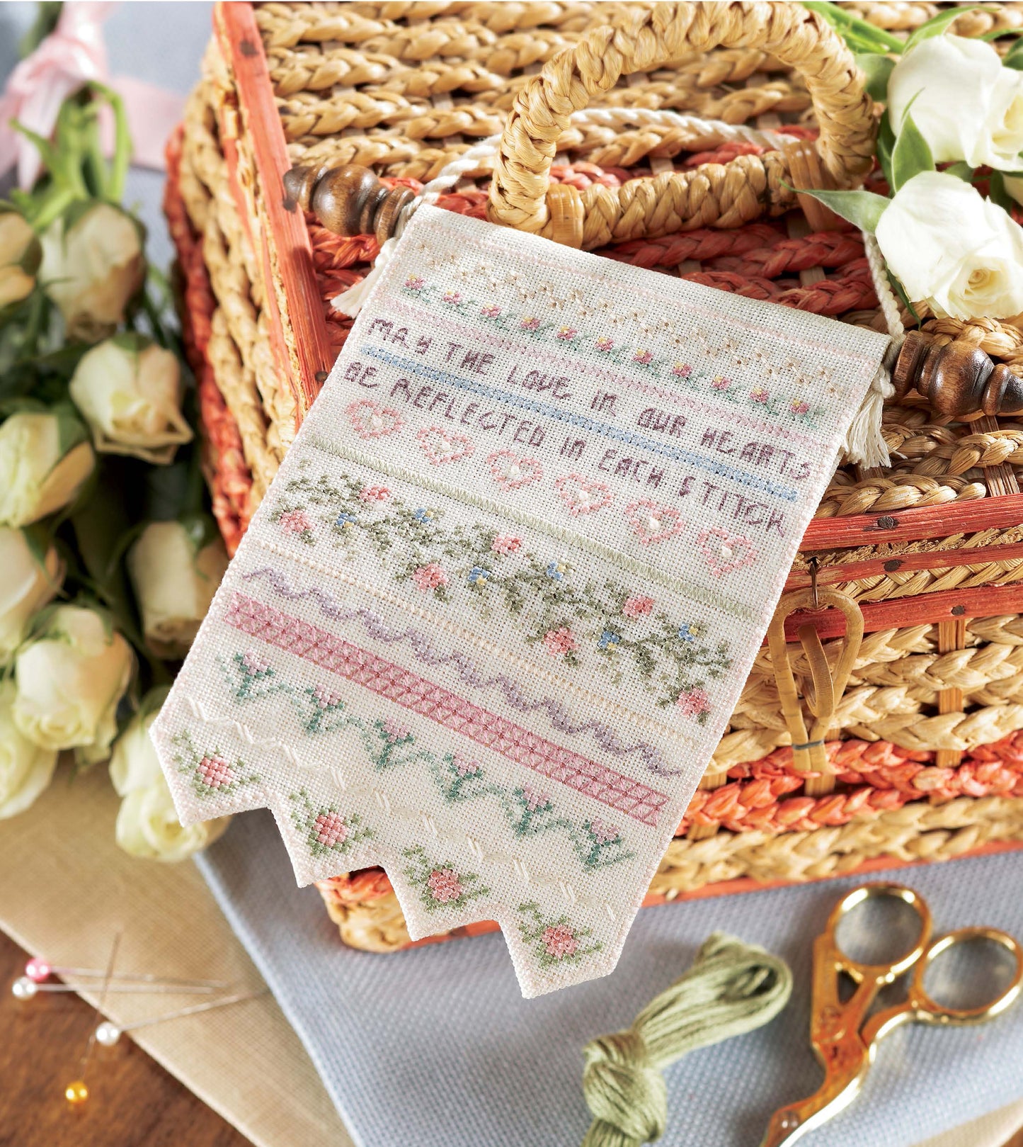 Samplers Cross Stitch by Leisure Arts features floral borders and the quote "May the love in our hearts be reflected in each stitch," shown with a wicker basket, white roses, scissors, and embroidery floss—ideal for sampler enthusiasts.