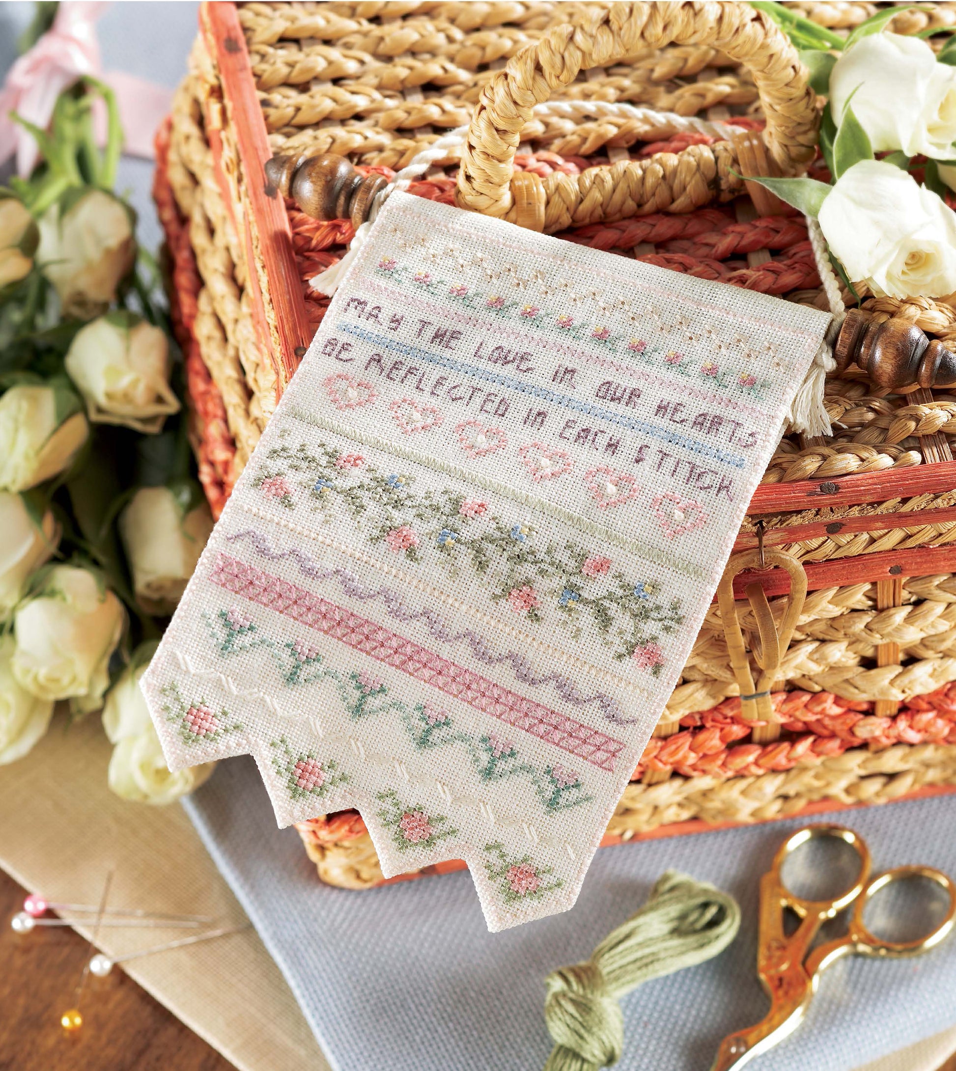 Samplers Cross Stitch by Leisure Arts features floral borders and the quote "May the love in our hearts be reflected in each stitch," shown with a wicker basket, white roses, scissors, and embroidery floss—ideal for sampler enthusiasts.