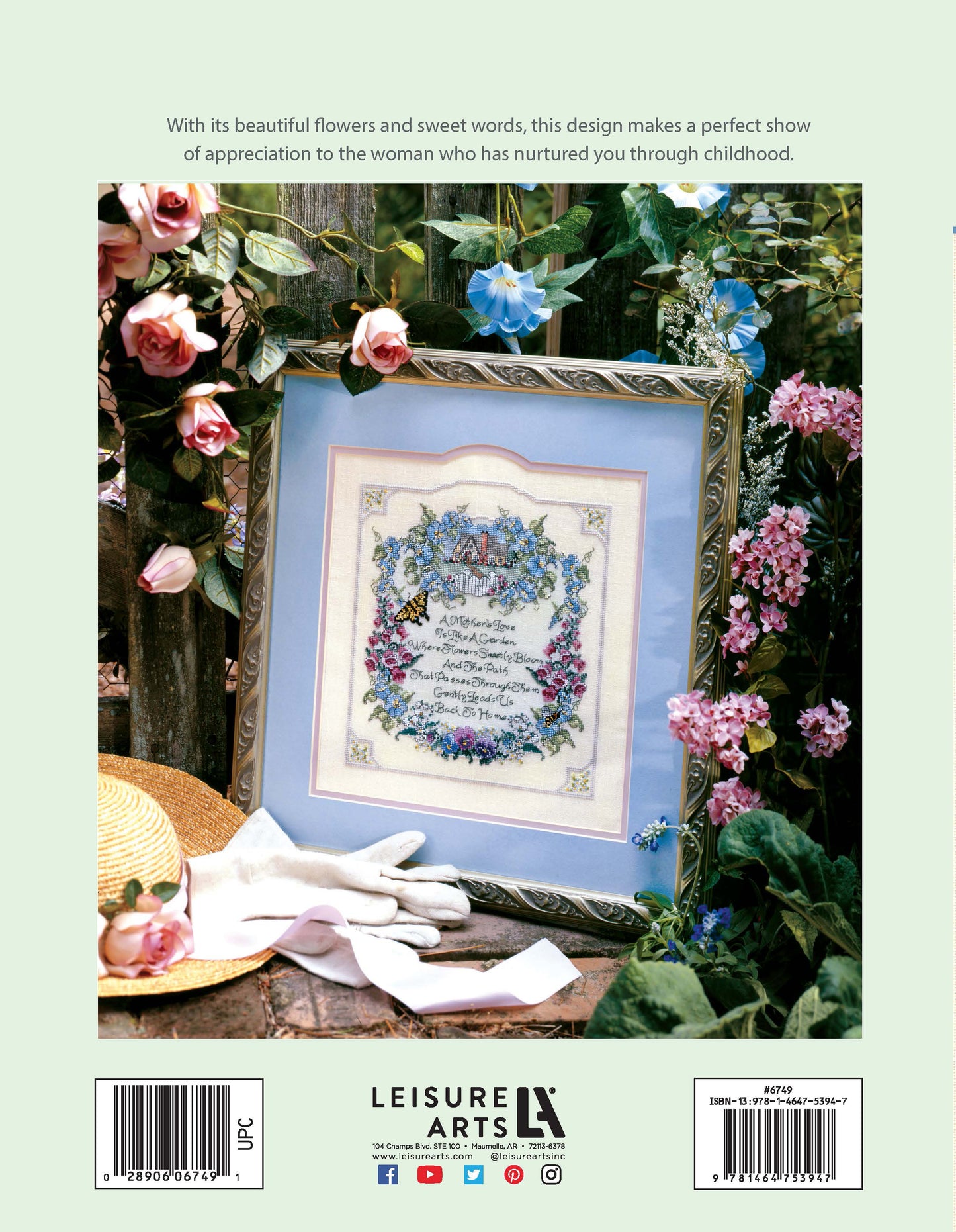 A Mother's Love by Leisure Arts—a framed floral cross stitch embroidery—rests on a garden chair among roses and greenery, with a straw hat and white gloves nearby, creating a peaceful vintage scene.