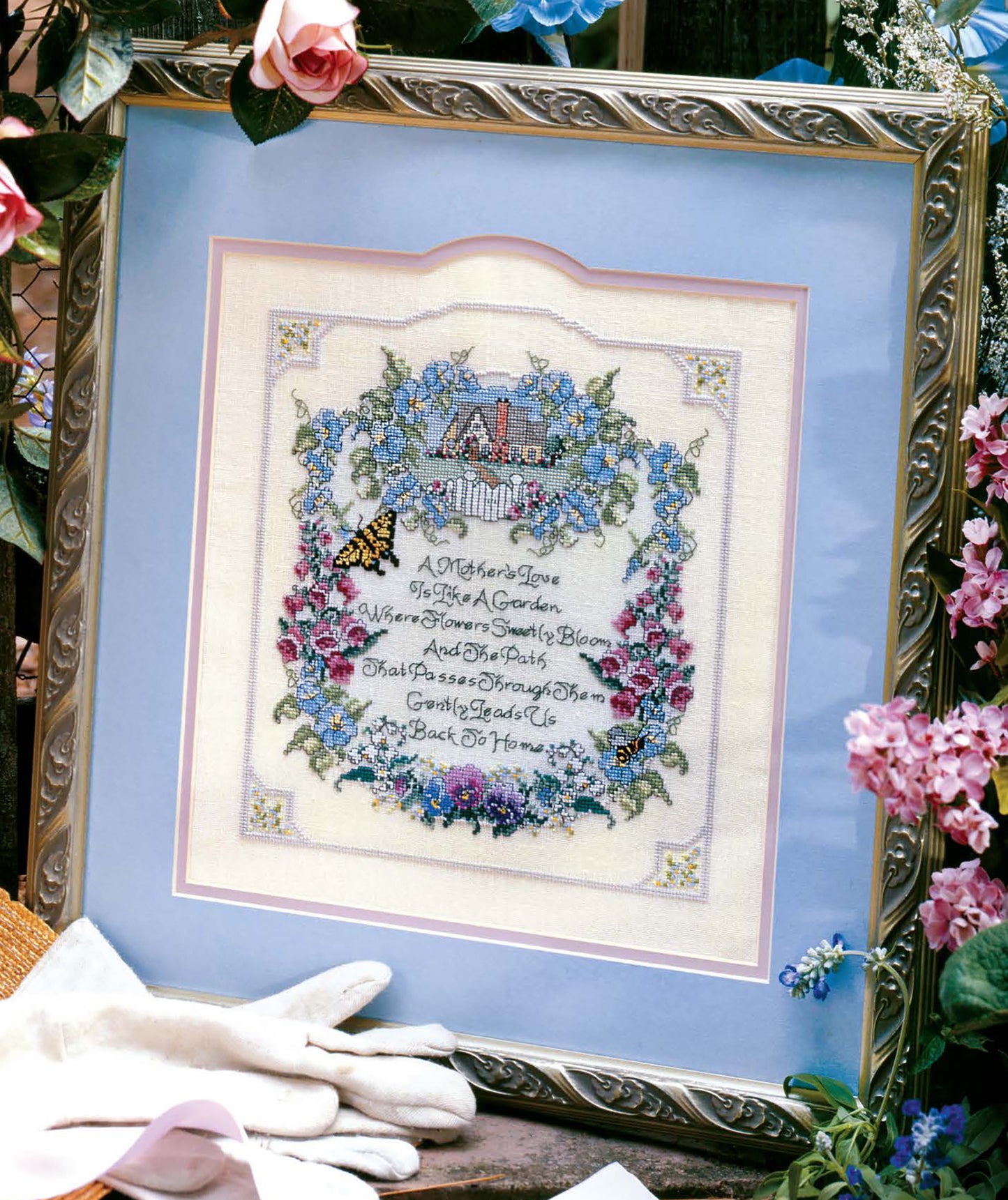 Leisure Arts’ “A Mother’s Love—Stitch a Beautiful Tribute to Motherly Love!” is a framed cross stitch with floral border, cottage, and butterfly, plus a loving quote surrounded by gardening gloves and flowers.
