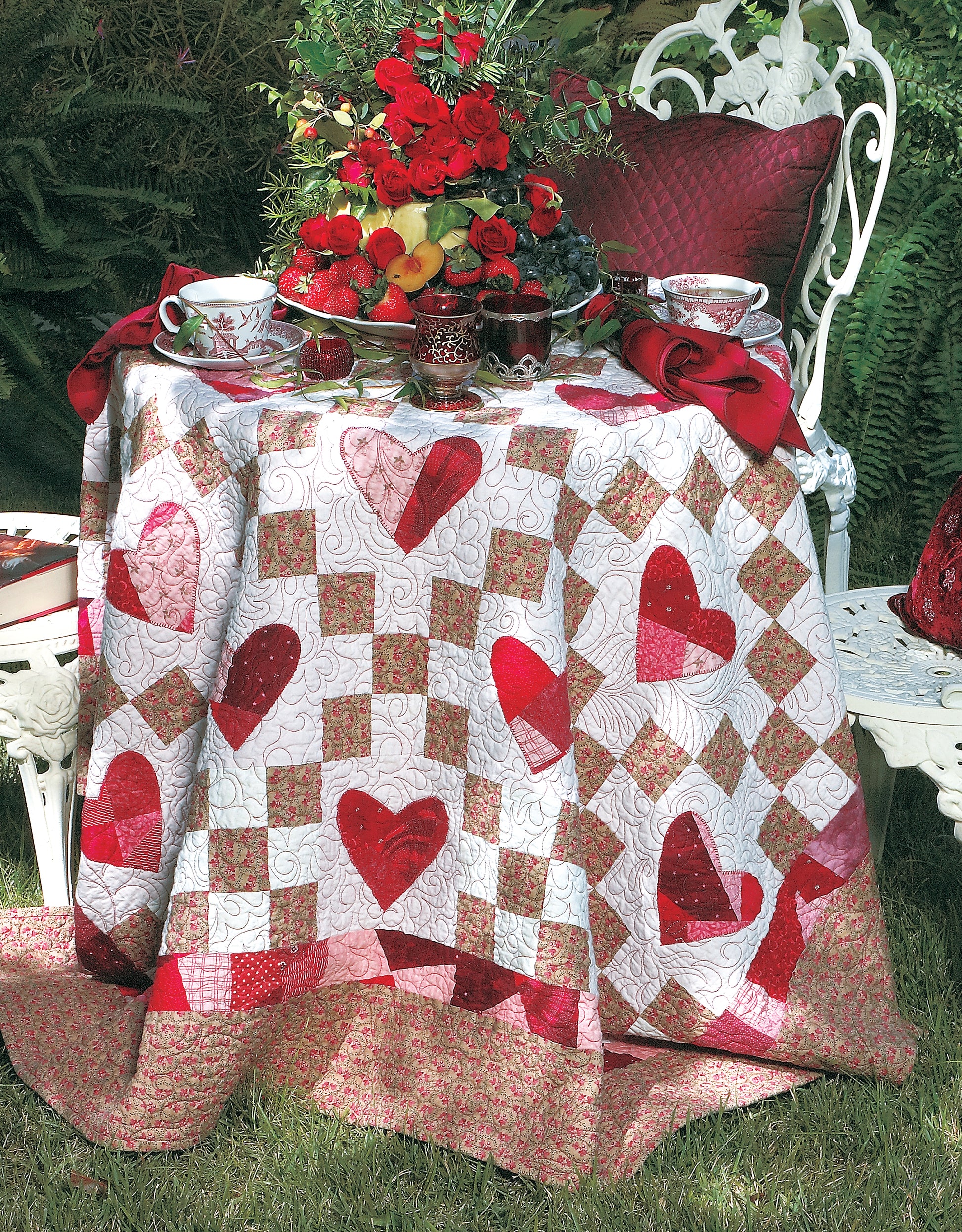 A decorative outdoor table is draped with Leisure Arts' Sentimental Hearts to Quilt, adorned with red hearts. Set with tea cups, fruit, and roses by a white metal chair on grass, it creates a romantic garden-inspired scene.