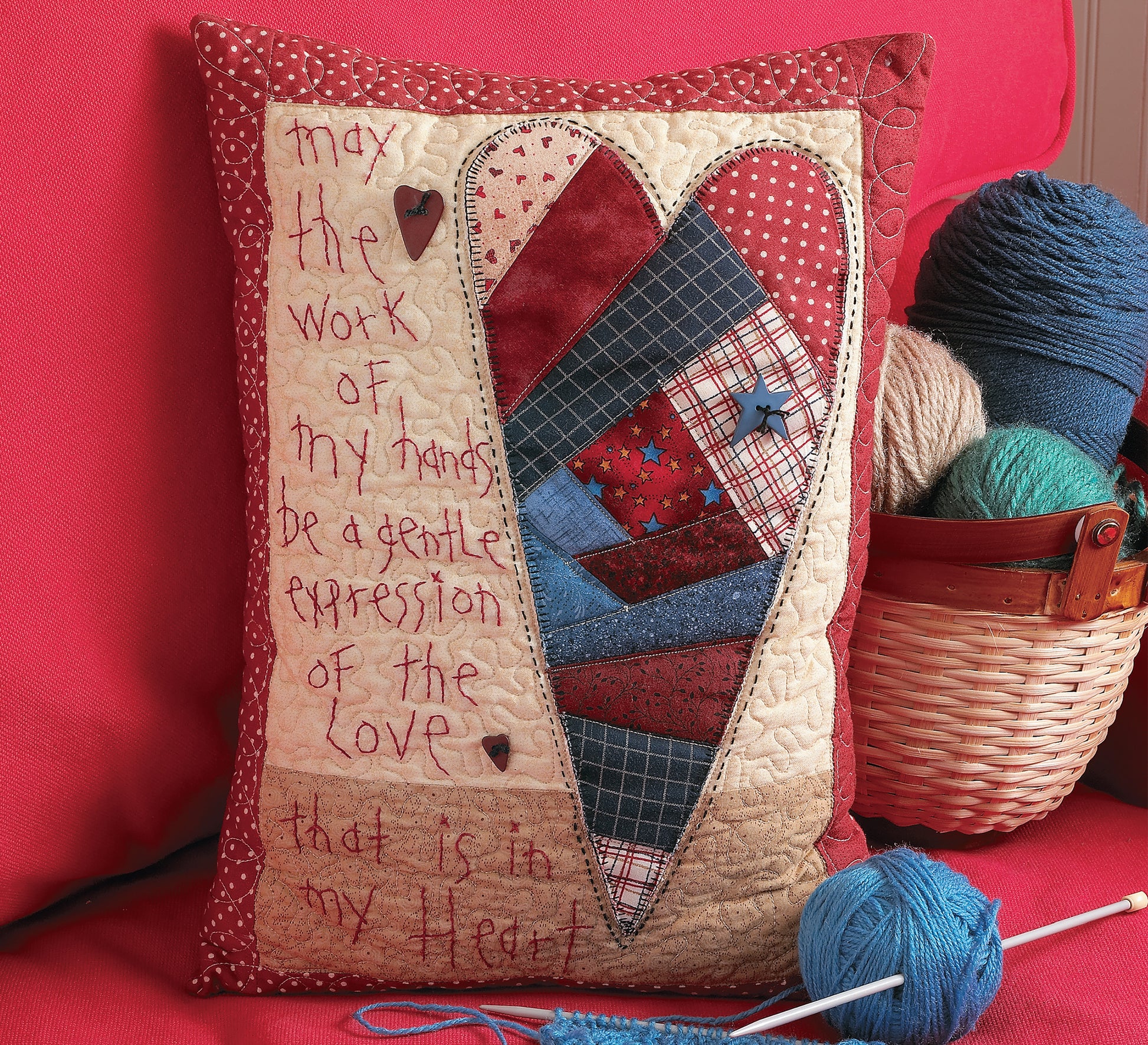 The Love & Friendship Quilted Pillows by Leisure Arts feature a patchwork heart and embroidered text, displayed on a red couch beside yarn, knitting needles, and a quilting book—inspiring heartfelt creativity in your home.