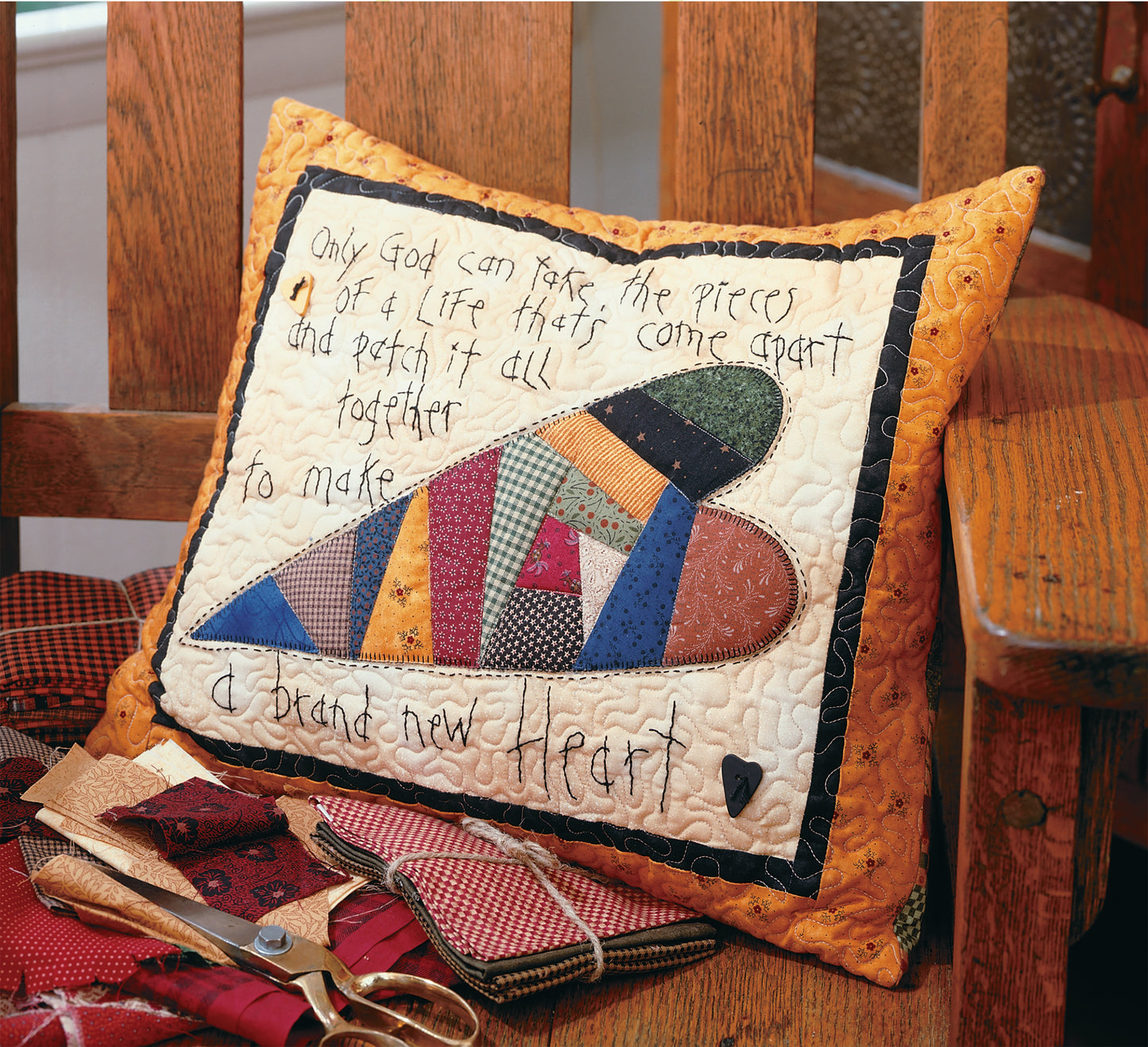 The Leisure Arts Love & Friendship Quilted Pillow, featuring a patchwork heart and uplifting message, rests on a wooden chair next to sewing supplies. The pillow’s text inspires: Only God can make a brand new heart from pieces of a life.