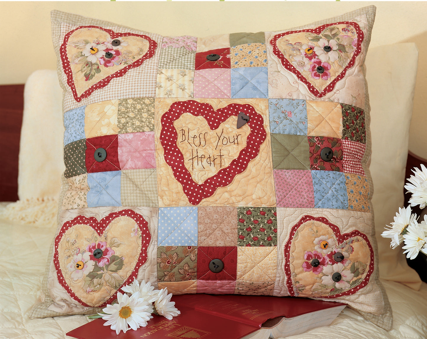 The Love & Friendship Quilted Pillows by Leisure Arts feature heart-shaped floral designs and "Bless Your Heart" embroidery, shown on a bed with daisies, a red book, and a quilting book—perfect inspiration for your sewing machine projects.