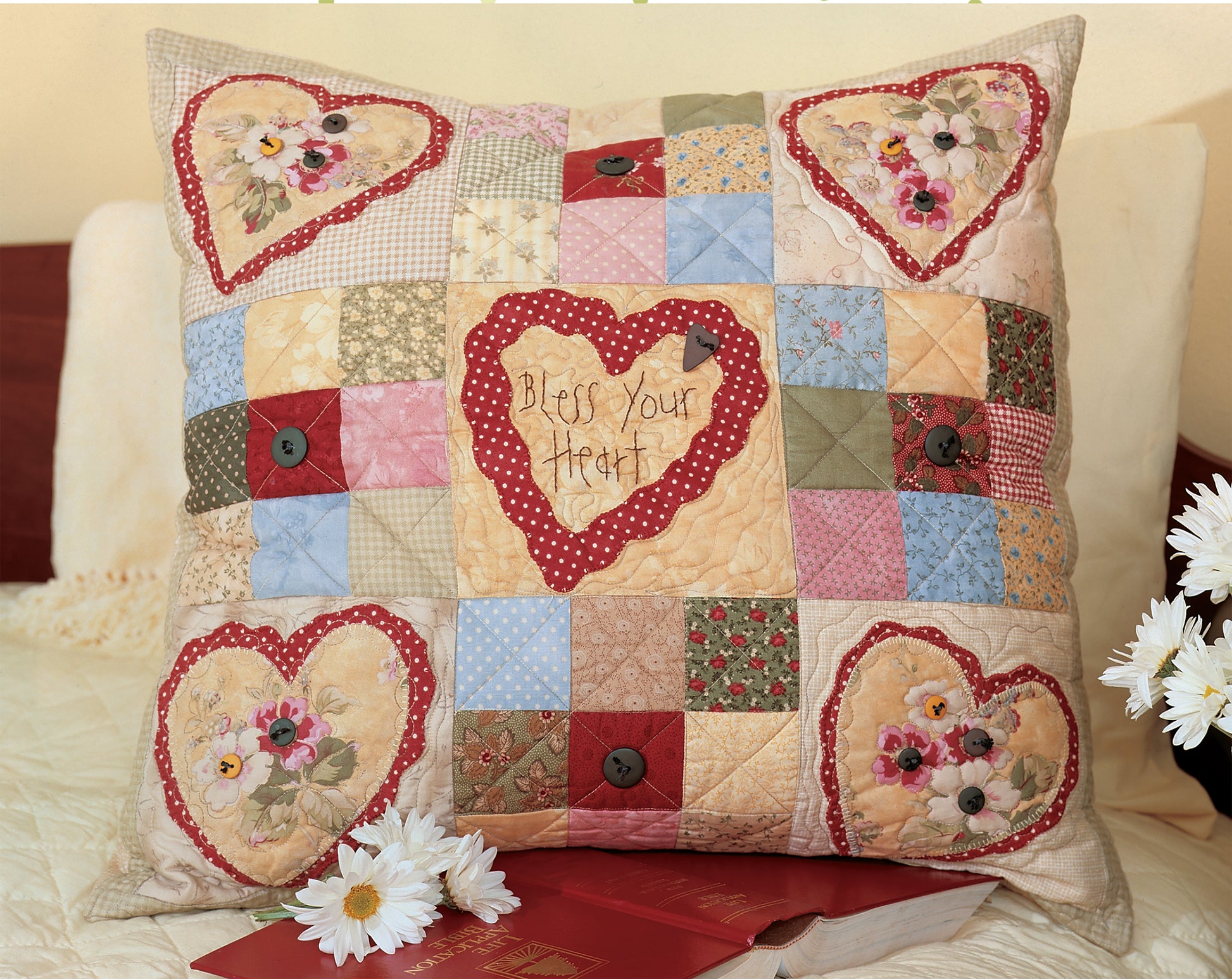 The Love & Friendship Quilted Pillows by Leisure Arts feature heart-shaped floral designs and "Bless Your Heart" embroidery, shown on a bed with daisies, a red book, and a quilting book—perfect inspiration for your sewing machine projects.