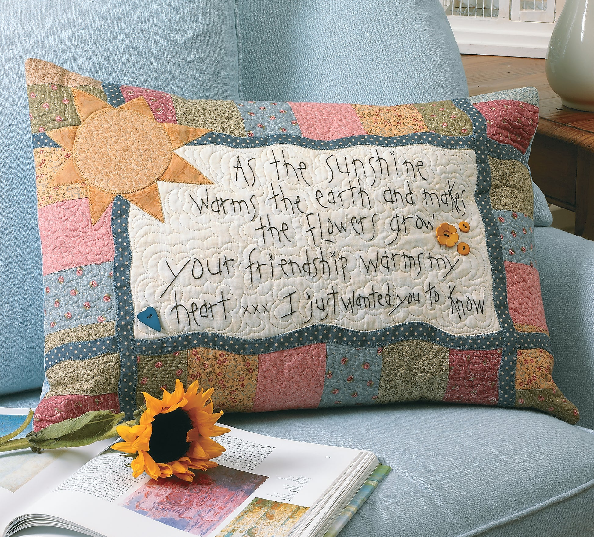 The Love & Friendship Quilted Pillow by Leisure Arts, crafted with free-motion quilting, features sun, fish, and flower designs plus friendship-themed embroidery. Shown on a blue chair near sunflowers and papers with sunlight streaming through a window.