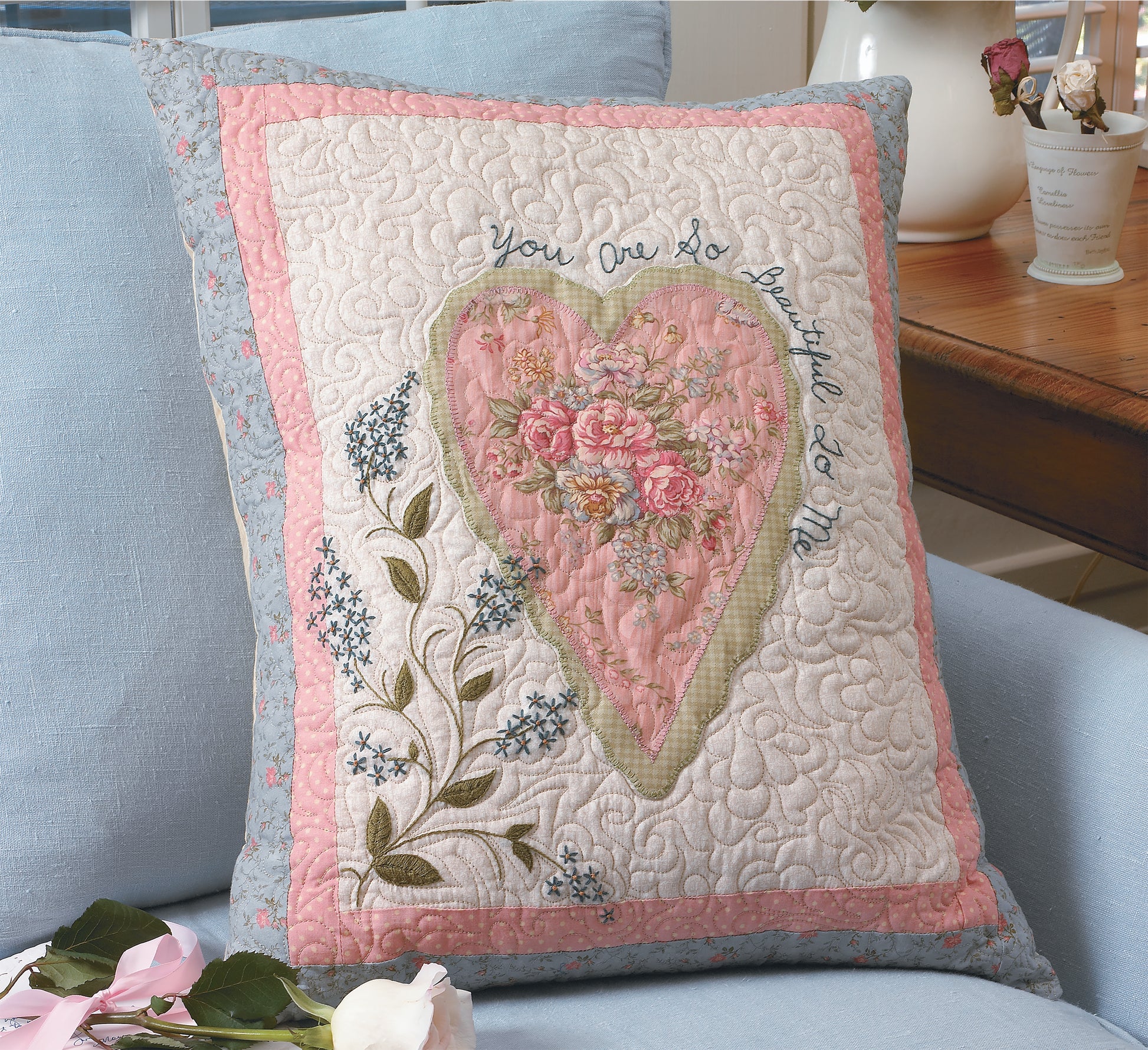 The Love & Friendship Quilted Pillow by Leisure Arts features a heart with embroidered pink flowers and green leaves, free-motion quilted, and the phrase "You are so beautiful to me" curved around it, shown on a light blue couch.
