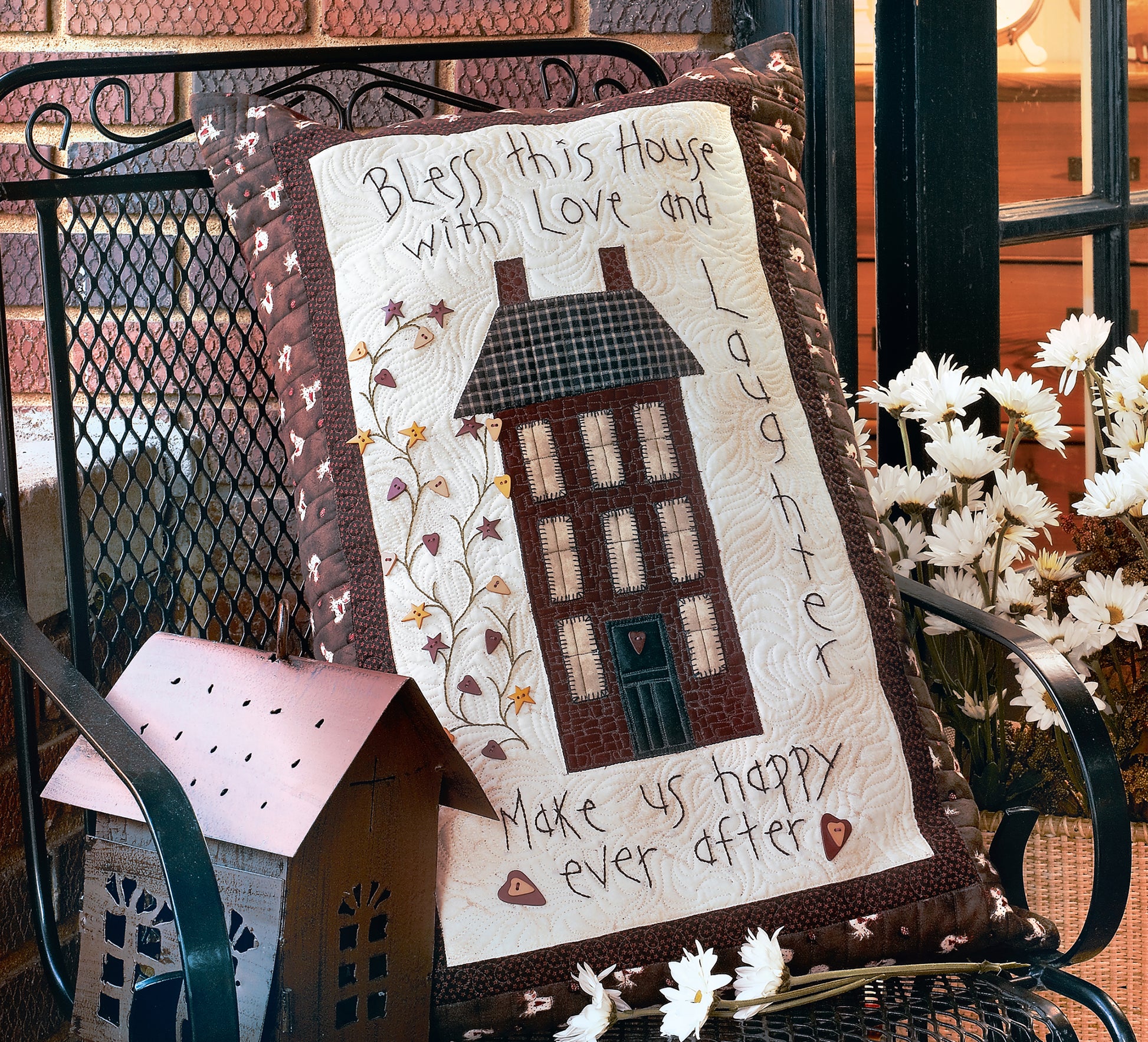 The Love & Friendship Quilted Pillows by Leisure Arts feature a charming embroidered house, flowers, and heartfelt words. Nearby are white daisies, a sewing machine, and a small house ornament in the foreground.
