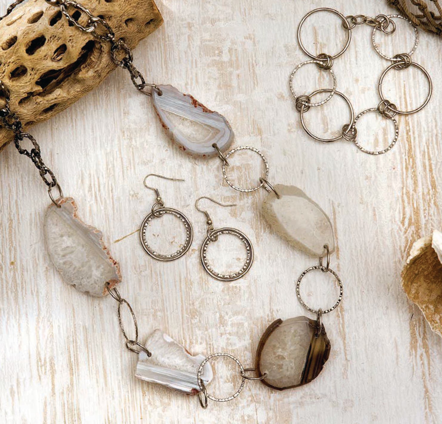 Wanderlust—Travel-Inspired Jewelry by Leisure Arts, featuring bold stone necklaces, eclectic earrings, and metal ring bracelets, is displayed on a light wooden surface with natural sponges for decoration.